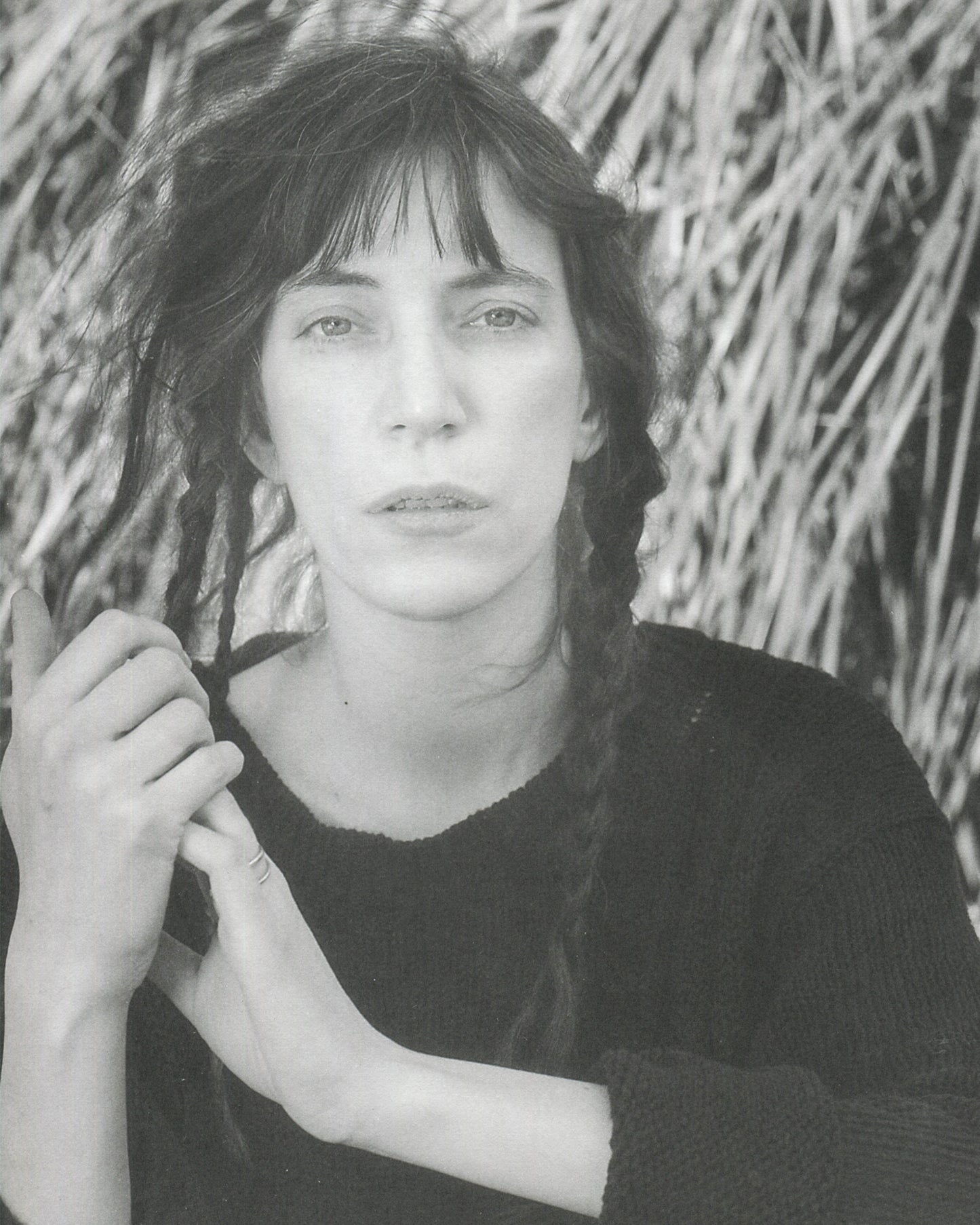 Interior page from Robert Mapplethorpe, Some Women, Bulfinch Press, 1992, showing a black-and-white portrait of Patti Smith, photographed outdoors against a background of dried straw or reeds, wearing a dark knitted sweater, her dark hair loose with a fringe, holding a braid of her hair in both hands, looking directly at the camera with a composed, direct expression.