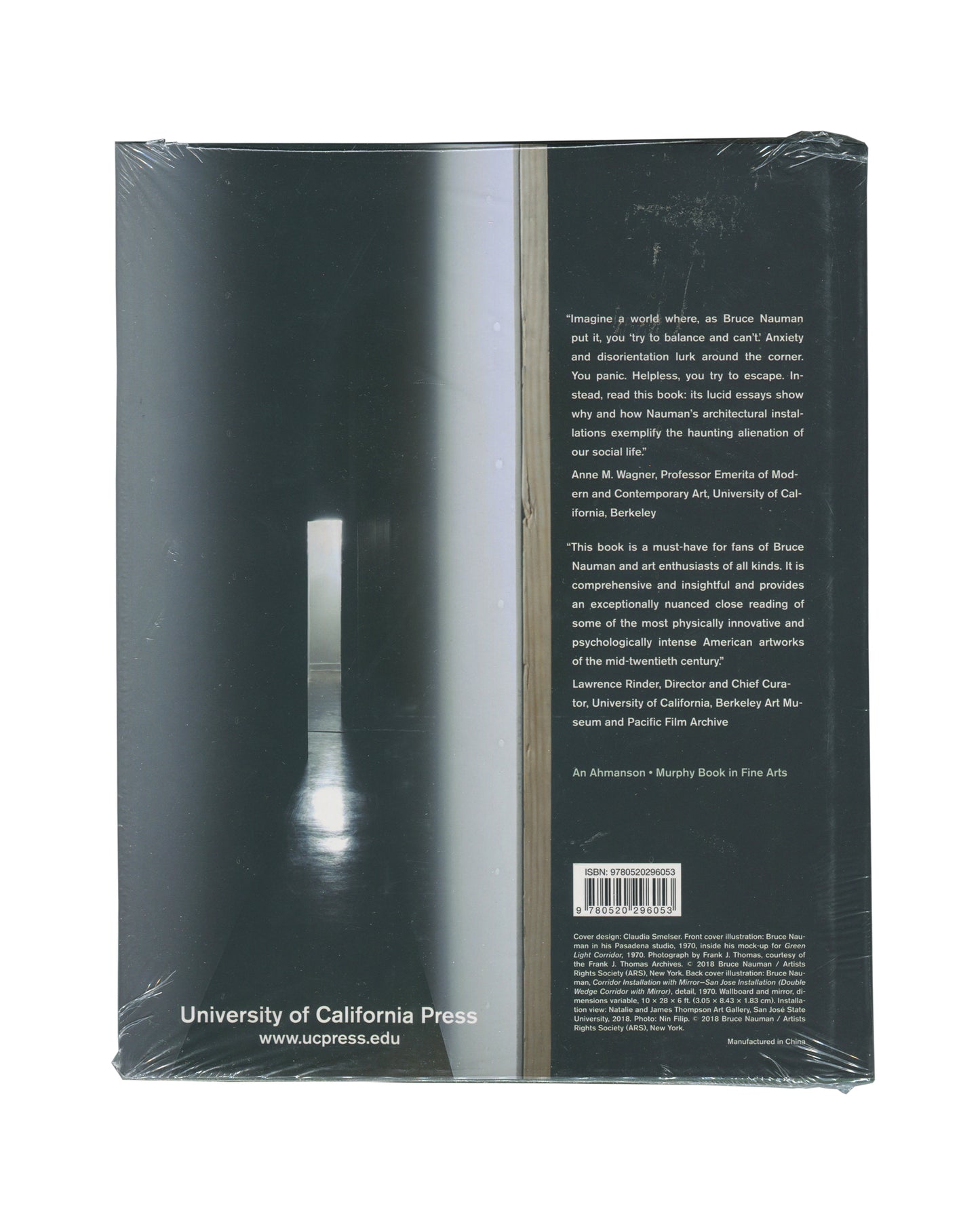 Monograph back cover, Bruce Nauman: Spatial Encounters, University of California Press, 2019. Sealed in distributor shrinkwrap. Dark teal-green ground. Full-bleed photograph — narrow corridor installation, single vertical strip of light at centre, reflective floor, deep spatial recession. Right column: two endorsement quotes — Anne M. Wagner, Professor Emerita of Modern and Contemporary Art, UC Berkeley; Lawrence Rinder, Director and Chief Curator, UC Berkeley Art Museum and Pacific Film Archive.