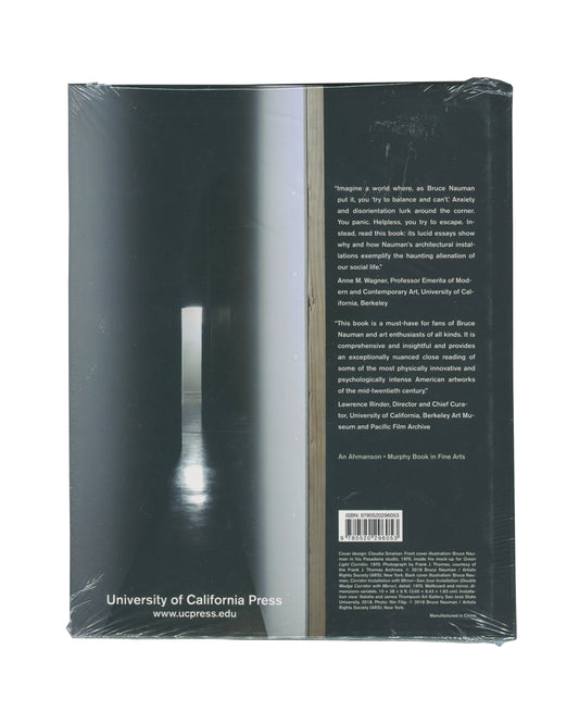 Monograph back cover, Bruce Nauman: Spatial Encounters, University of California Press, 2019. Sealed in distributor shrinkwrap. Dark teal-green ground. Full-bleed photograph — narrow corridor installation, single vertical strip of light at centre, reflective floor, deep spatial recession. Right column: two endorsement quotes — Anne M. Wagner, Professor Emerita of Modern and Contemporary Art, UC Berkeley; Lawrence Rinder, Director and Chief Curator, UC Berkeley Art Museum and Pacific Film Archive.