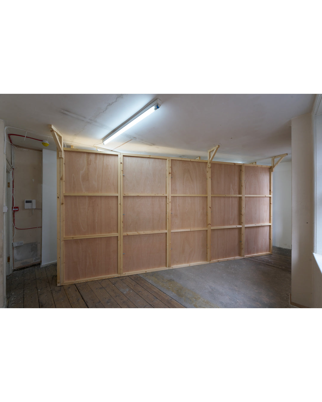 Installation view, THE NEW RARE EDITIONS, 2026. Full-width pine modular wall structure spanning the room; fluorescent tube overhead; worn wooden floorboards; door and electrical panel visible at left.