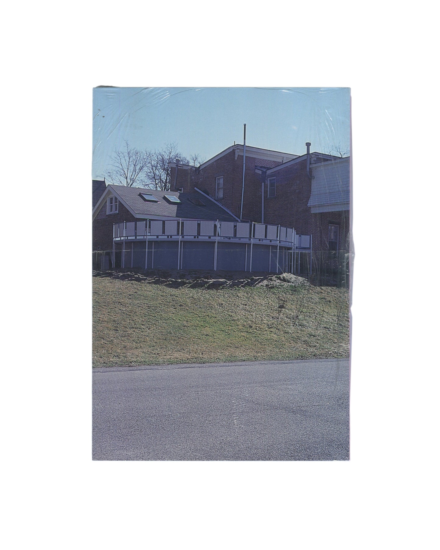 Rear cover of Richard Prince’s The Girl Next Door, sealed in original shrinkwrap, showing a suburban house, deck, lawn, and road photographed in muted daylight.