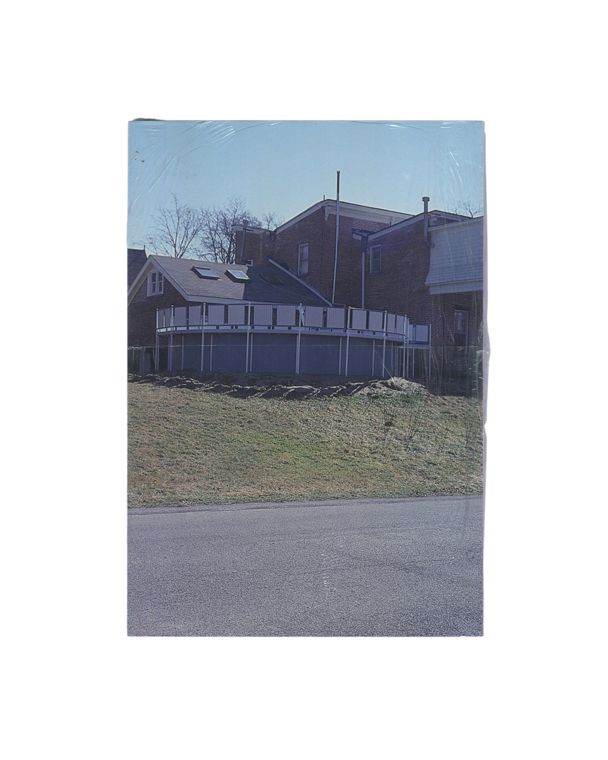 Rear cover of Richard Prince’s The Girl Next Door, sealed in original shrinkwrap, showing a suburban house, deck, lawn, and road photographed in muted daylight.