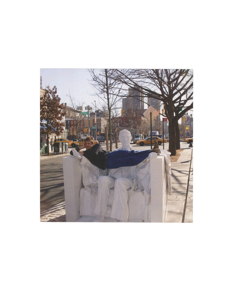 Original 2009 Adel Abdessemed Rio exhibition invitation card, David Zwirner, New York — recto reproducing a detail from Lincoln, 2009, C-print, 50 × 68½ inches: colour photograph of the artist reclining across a seated white sculptural figure based on the Lincoln Memorial, shot on a New York street corner in winter light with bare trees, yellow taxis, and urban buildings visible in background.