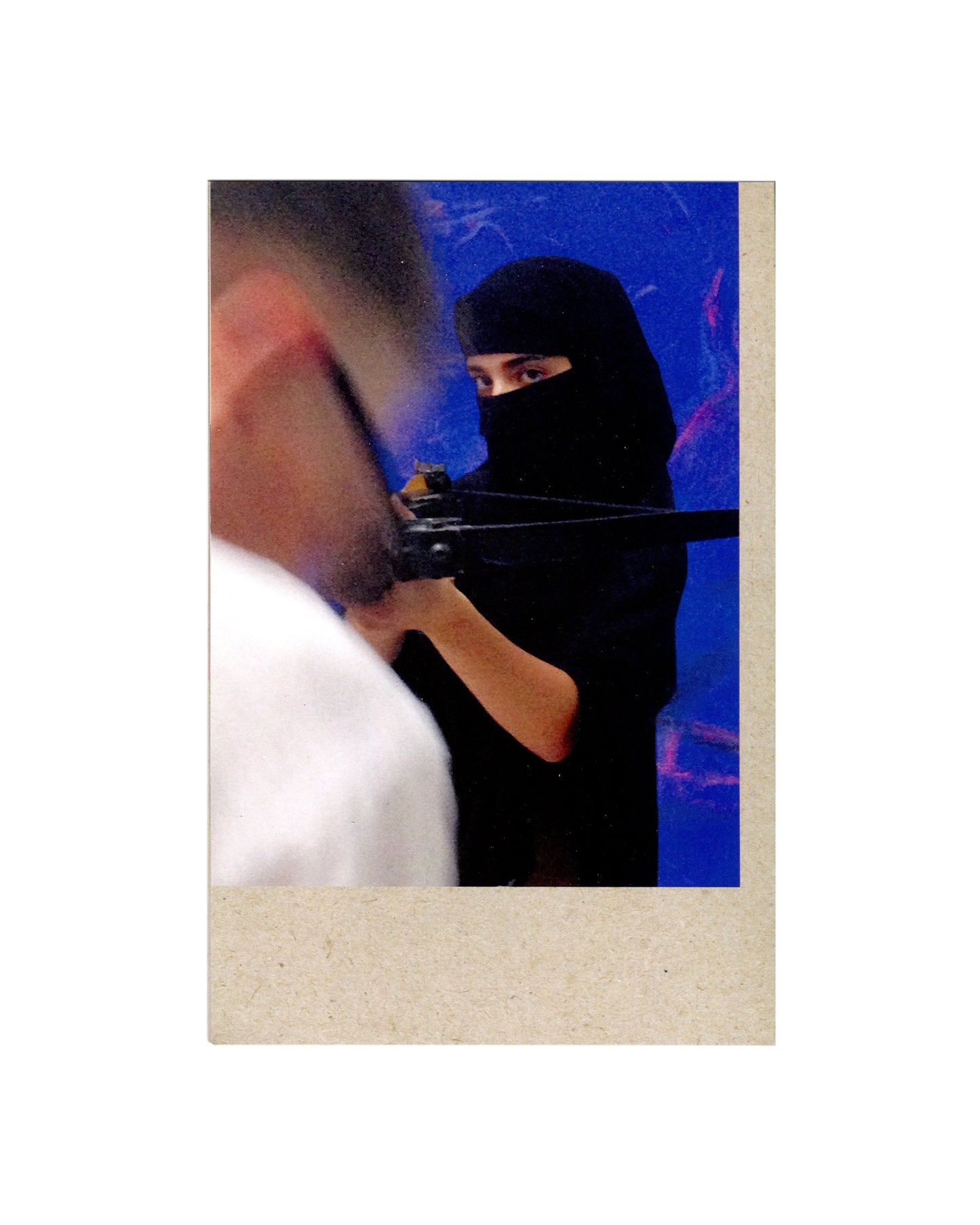 Interior spread of 2016 Rita Ackermann Jezebels showing a colour photograph of a masked woman in black niqab aiming a crossbow at a man in a white shirt in the foreground, against a blue painted ground by Ackermann.