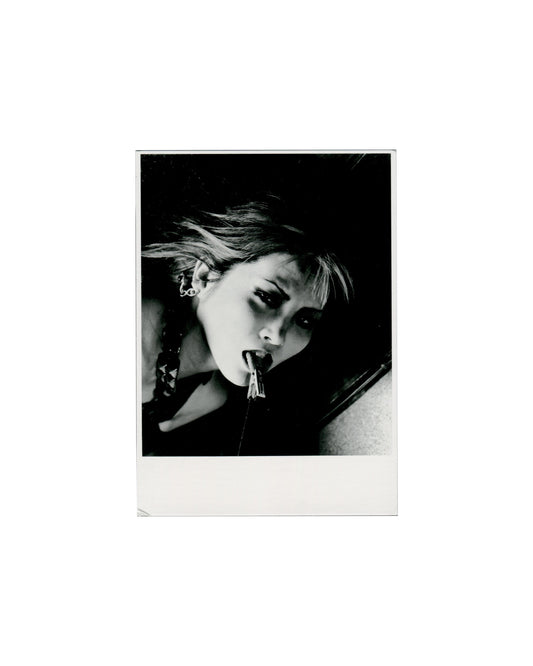 Recto — photograph Nobuyoshi Araki, Untitled, 1995, Tokyo Novelle, Kunstmuseum Wolfsburg, 1995–96. Artist postcard recto: monochrome photograph within a Polaroid-like white border — close-up of a young woman's face tilted sideways against a dark background, hair loose and wind-blown, a small clothes peg held between her lips, hoop earring visible, braided hair at left; high-contrast black-and-white printing. 10.5 × 15 cm.

