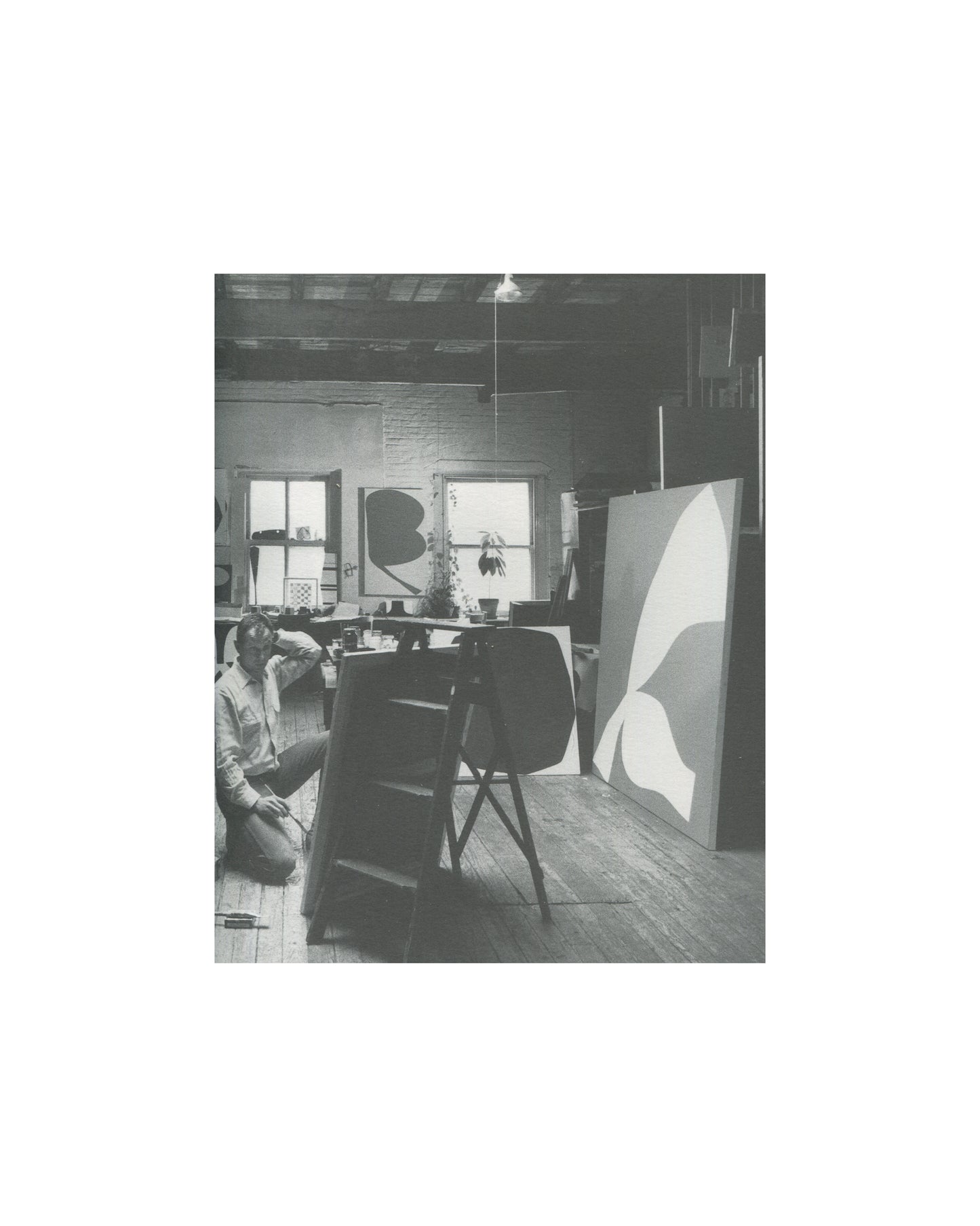 Ellsworth Kelly — A Salute to Ellsworth Kelly, 1999 — interior plate. Black and white photograph of Ellsworth Kelly seated on the floor of his studio, working at low level. Two large canvases with biomorphic curved forms visible in background. Worktable and studio equipment in mid-ground.