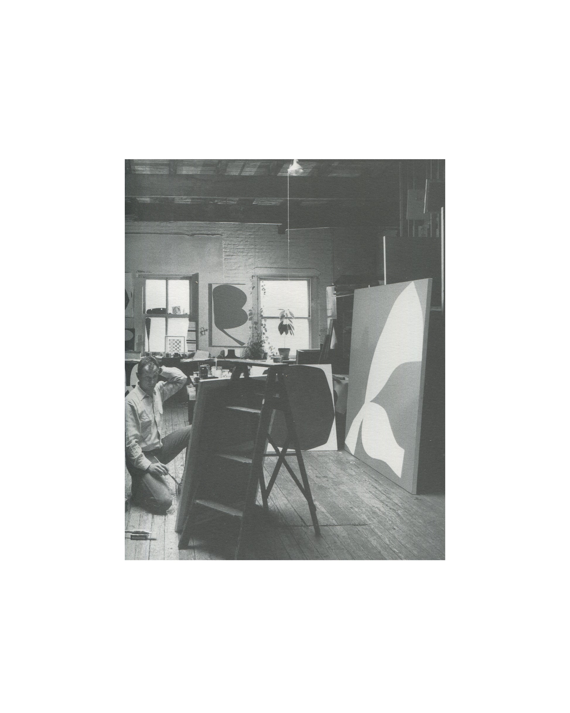Ellsworth Kelly — A Salute to Ellsworth Kelly, 1999 — interior plate. Black and white photograph of Ellsworth Kelly seated on the floor of his studio, working at low level. Two large canvases with biomorphic curved forms visible in background. Worktable and studio equipment in mid-ground.