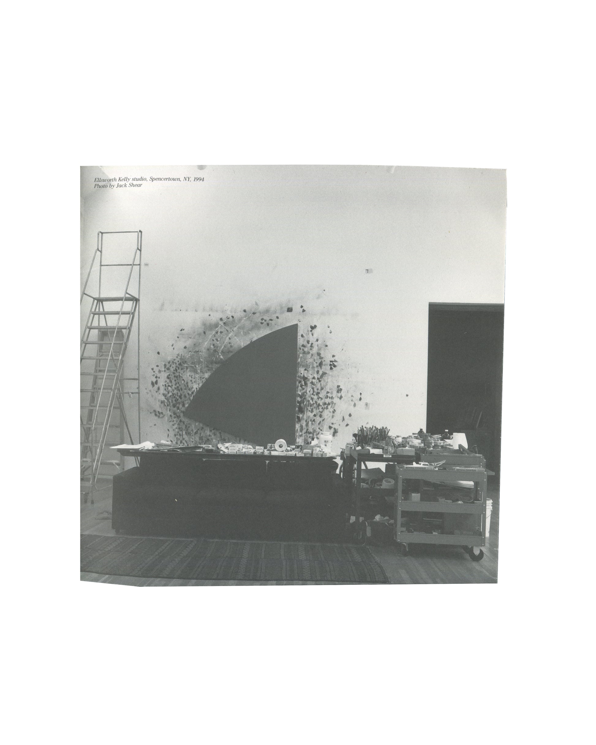 Ellsworth Kelly — A Salute to Ellsworth Kelly, 1999 — interior plate. Black and white photograph of Kelly's studio, Spencertown, NY, 1994. Photo by Jack Shear. Large dark quarter-circle canvas installed against white wall, surrounded by scattered paper studies. Metal stepladder left, worktable and trolley in foreground.