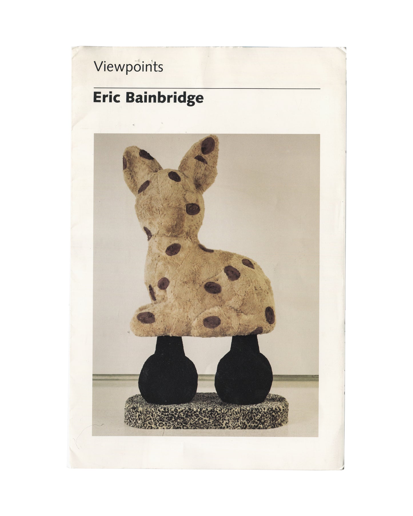 Original 1986 Eric Bainbridge Viewpoints Walker Art Center Minneapolis exhibition brochure cover showing white ground with 'Viewpoints' in small light serif type at upper left, 'Eric Bainbridge' in bold black serif type below a thin green rule, and a colour photograph of Bainbridge's sculpture Handle (1986): a large seated rabbit-like form covered in beige fur fabric with dark brown oval spots, mounted on two black rounded leg forms on a grey speckled oval base, photographed from behind against a white gall