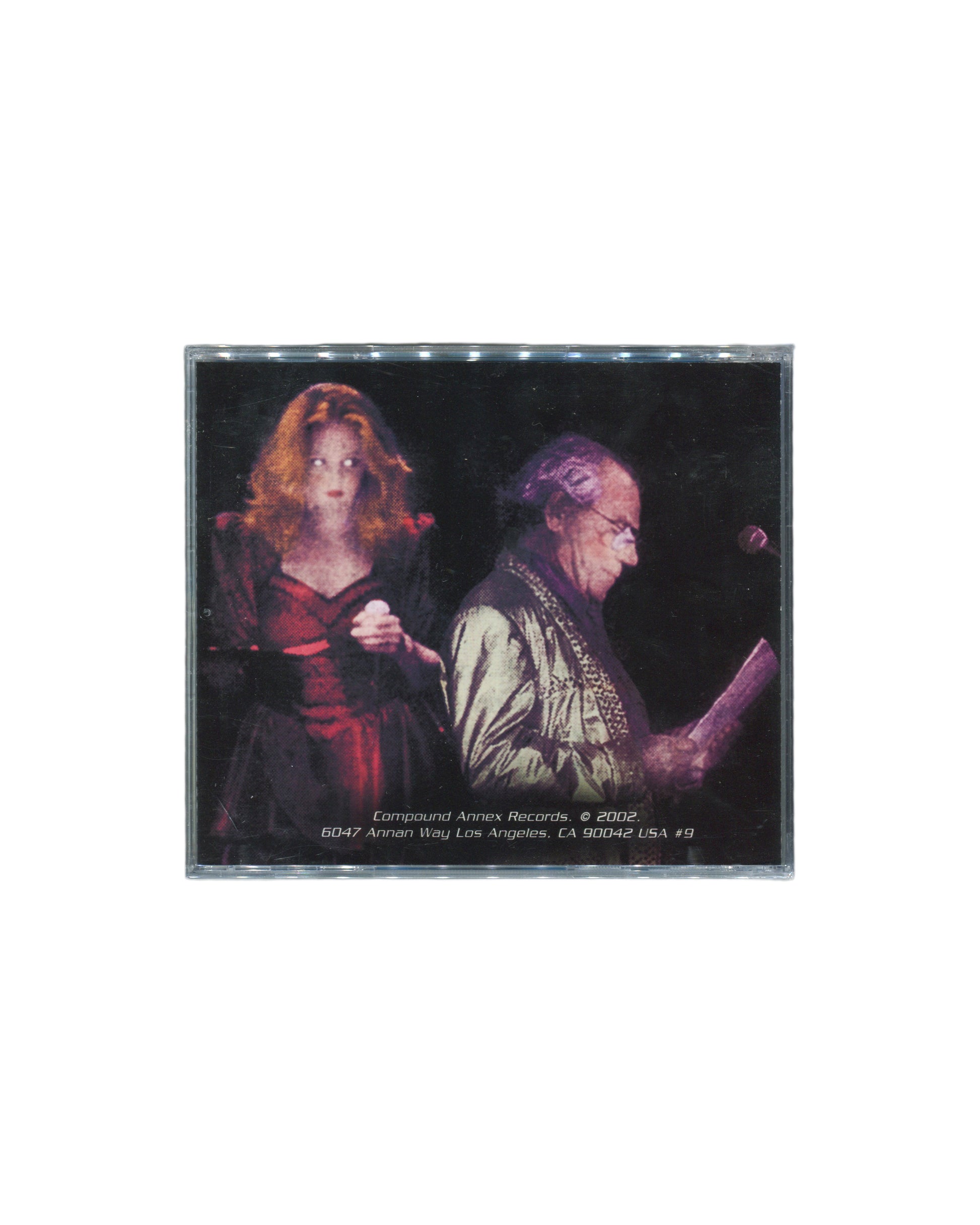 Reverse of 2002 Jean Baudrillard & The Chance Band Suicide Moi CD, Compound Annex Records — dark back cover with colour photograph of two performers on stage, a woman in a red dress at left and an older man reading from papers at a microphone at right, Compound Annex Records copyright line and Los Angeles address at foot

