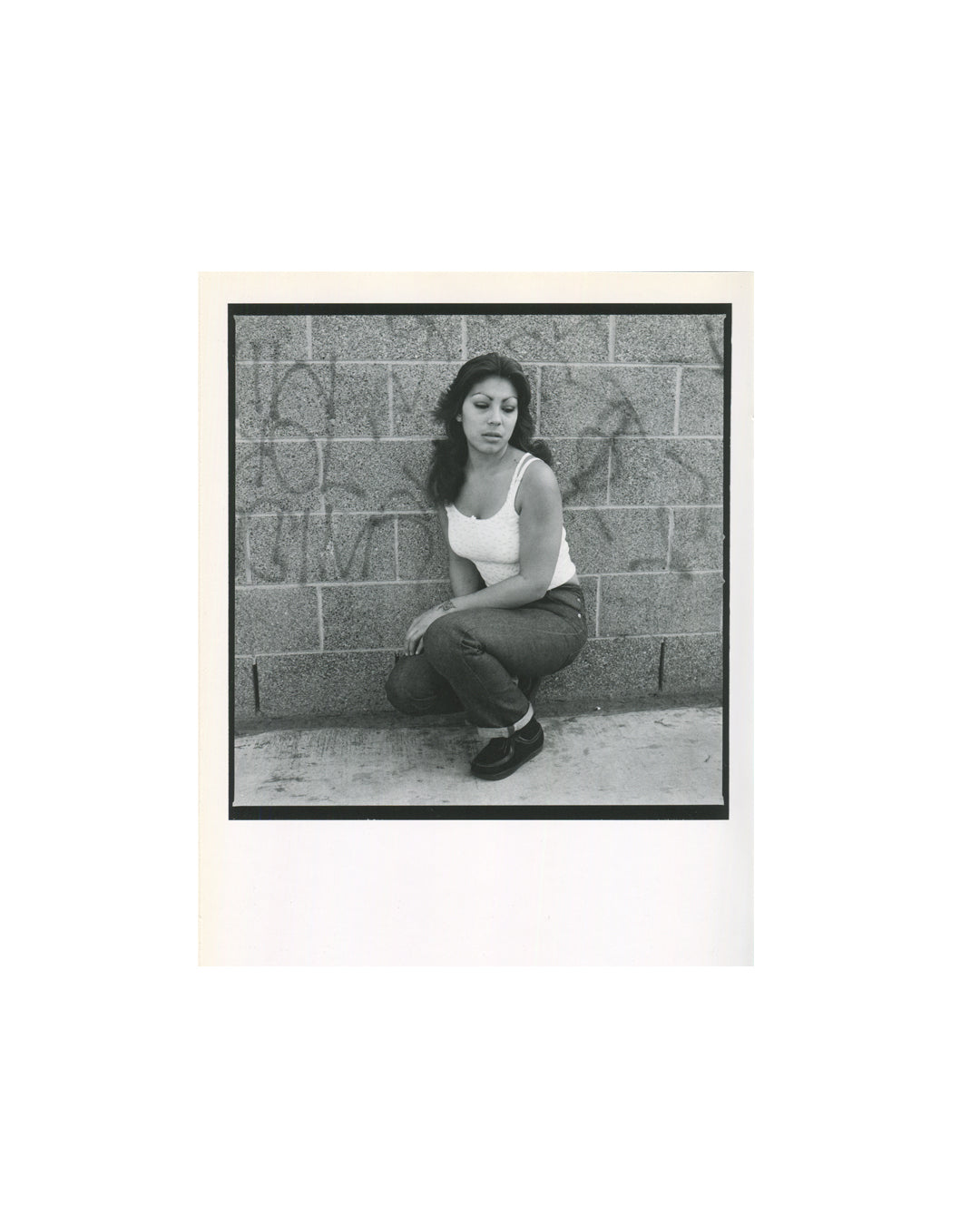 Interior spread showing black-bordered portrait of a young woman crouching against a graffiti-covered cinder block wall, wearing a white tank top and dark jeans.