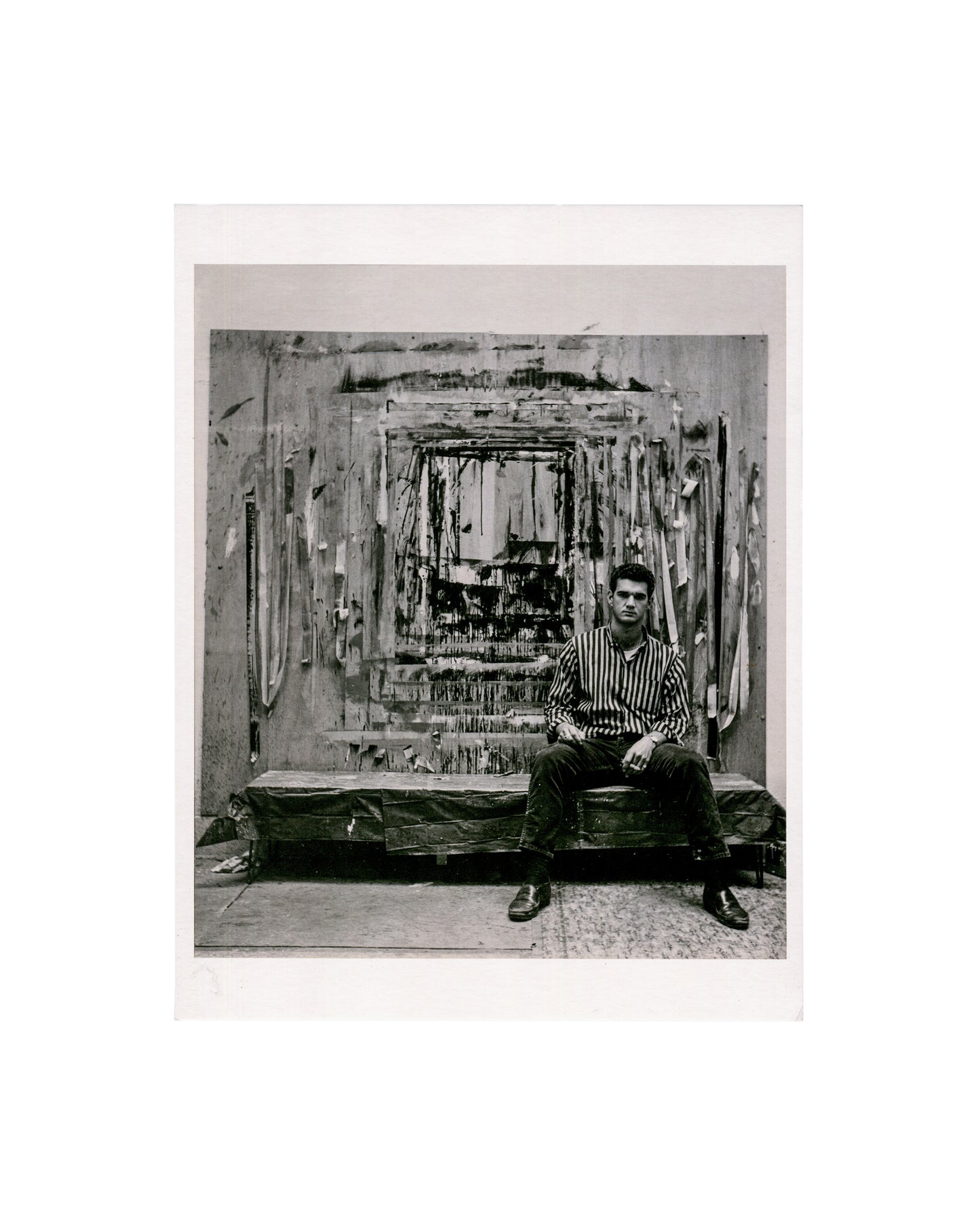 Larry Bell in his Marine Street studio, Venice, California, c.1959, photograph by Jerry McMillan; black and white, Bell seated before large mixed-media wall work
