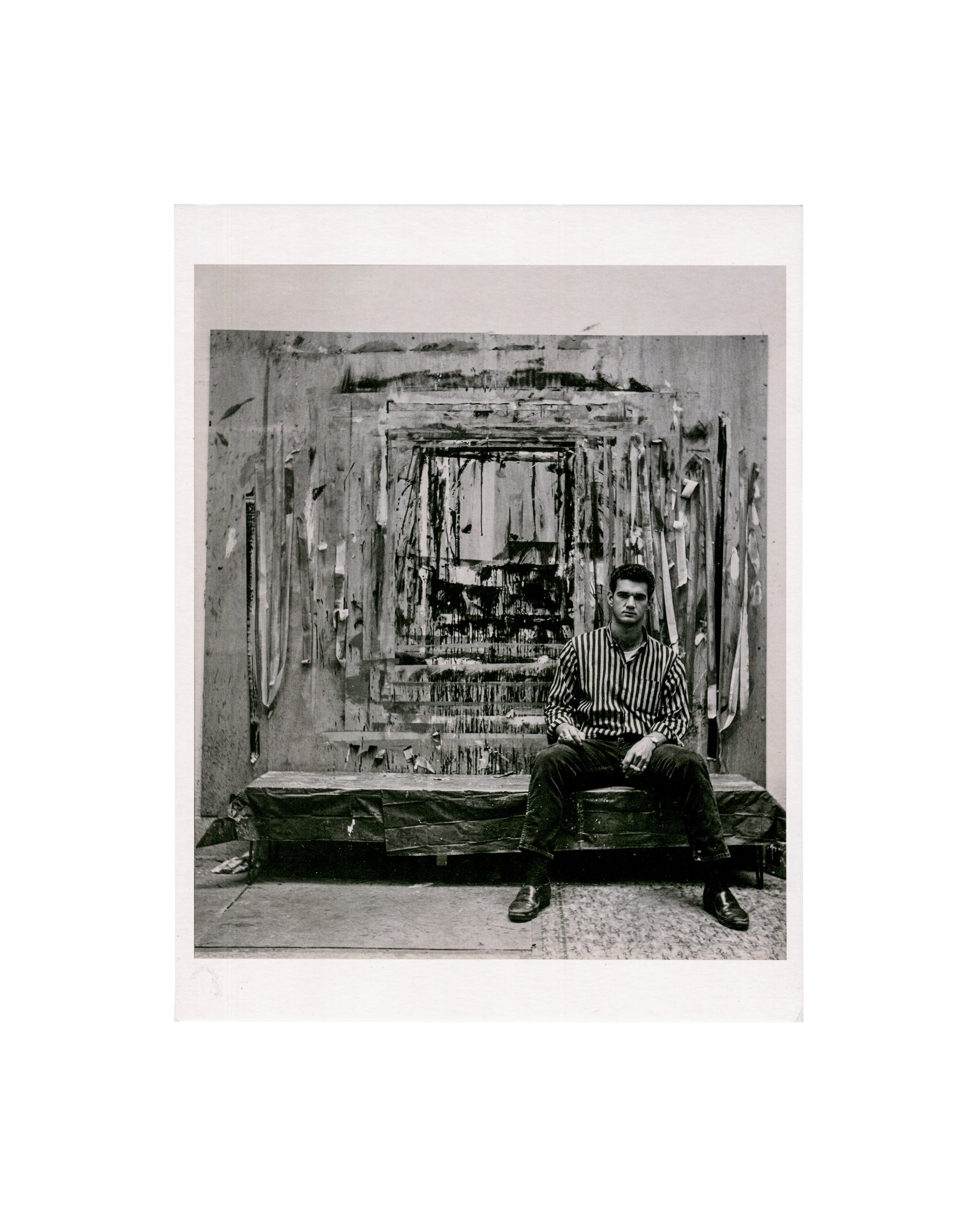 Larry Bell in his Marine Street studio, Venice, California, c.1959, photograph by Jerry McMillan; black and white, Bell seated before large mixed-media wall work
