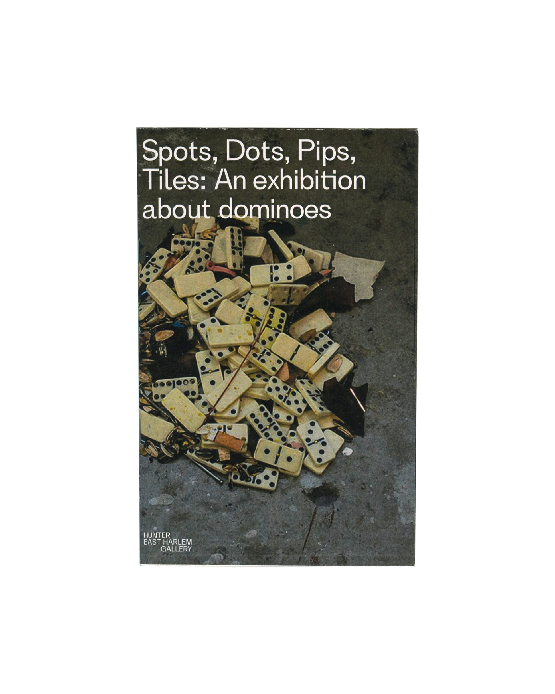 Original 2016 Spots, Dots, Pips, Tiles: An Exhibition about Dominoes exhibition catalogue, Hunter East Harlem Gallery, Hunter College, New York. Front cover showing a narrow vertical format with a split design: left half white with the exhibition title in stacked bold sans-serif type, right half a cropped colour photograph of scattered domino tiles and debris on grey pavement. 13 × 20.5 cm, staple-bound softcover.