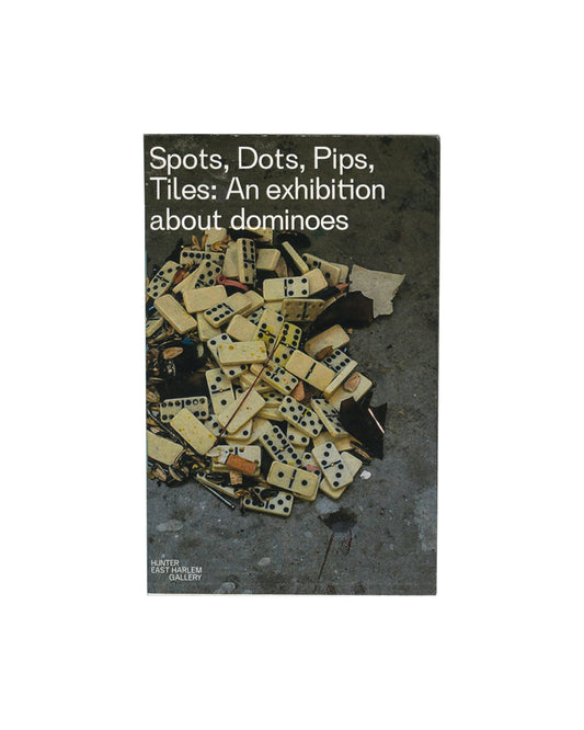 Original 2016 Spots, Dots, Pips, Tiles: An Exhibition about Dominoes exhibition catalogue, Hunter East Harlem Gallery, Hunter College, New York. Front cover showing a narrow vertical format with a split design: left half white with the exhibition title in stacked bold sans-serif type, right half a cropped colour photograph of scattered domino tiles and debris on grey pavement. 13 × 20.5 cm, staple-bound softcover.