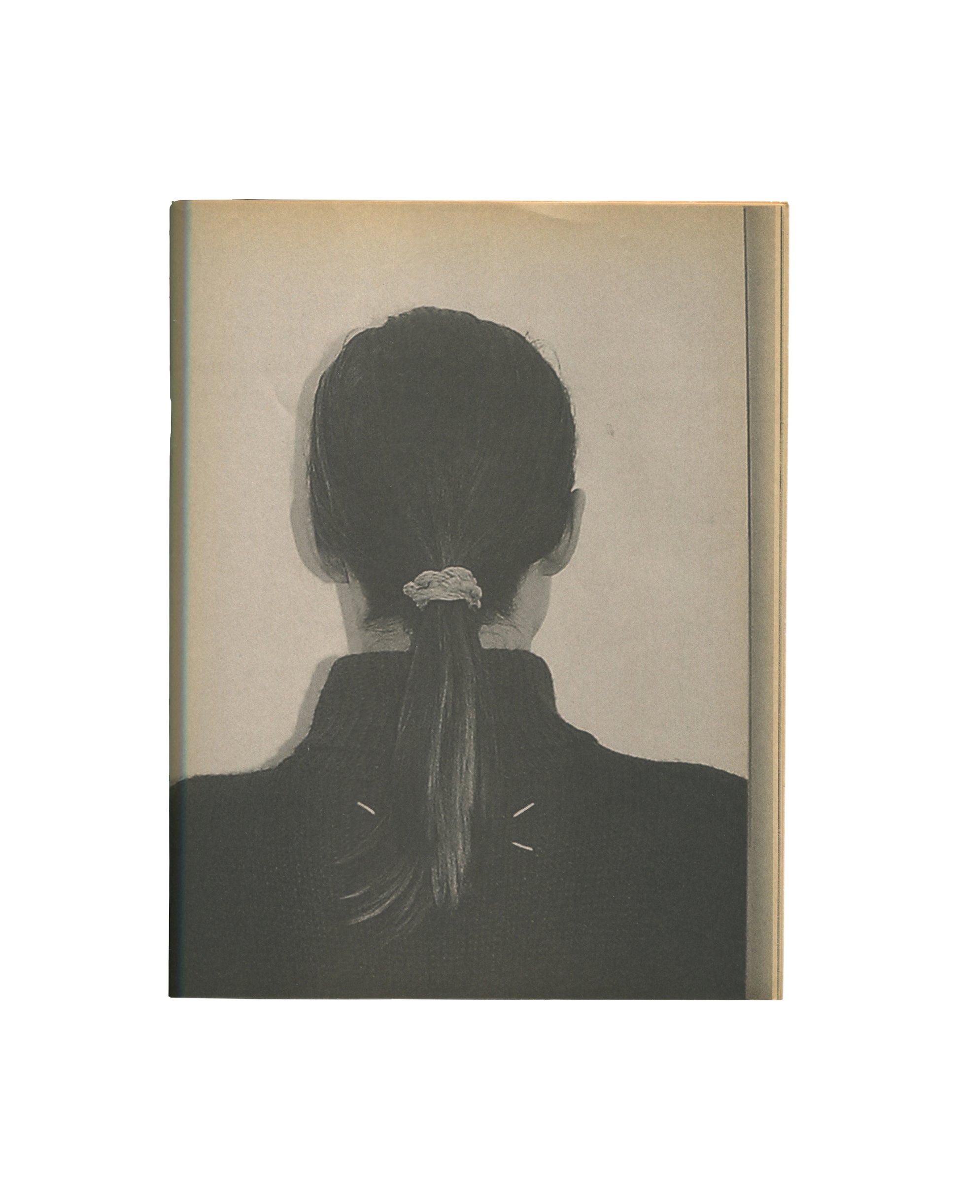 Interior page of c. 2002 Mark Borthwick Xerox edition of 2000-1, Maison Martin Margiela. Single page showing a grayscale Xerox photograph of the back of a woman's head and upper back: dark hair pulled into a low ponytail secured with a pale scrunchie, wearing a dark high-necked top with two small diagonal pin marks visible at the upper back. Shot against a plain pale ground. No caption or text. Copier grain and tonal softening visible.

