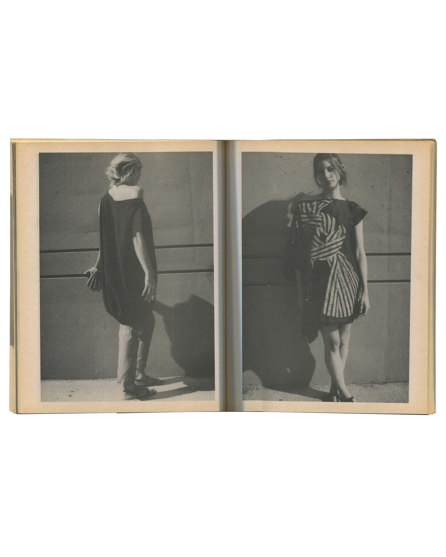 Interior spread of c. 2002 Mark Borthwick Social Documentaries Amid this Piste, self-published photocopy edition. Double-page spread showing two grayscale Xerox fashion photographs printed full-bleed across both pages: left image showing a woman from behind in a plain dark short-sleeved shift dress, standing against a pale wall with a strong cast shadow, wearing heels; right image showing the same woman facing three-quarters toward the camera in a bold black-and-white diagonal striped wrap dress with flutte