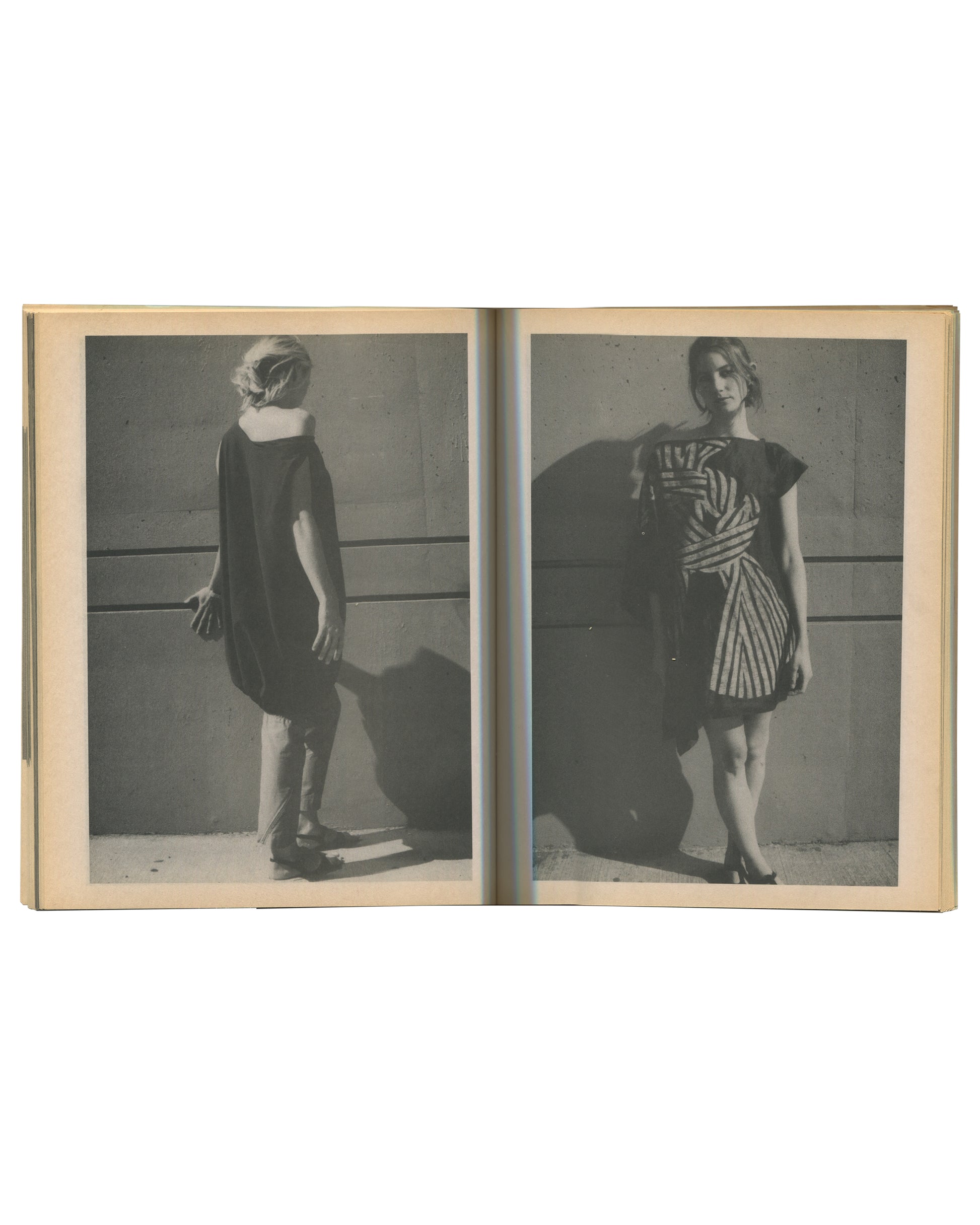 Interior spread of c. 2002 Mark Borthwick Social Documentaries Amid this Piste, self-published photocopy edition. Double-page spread showing two grayscale Xerox fashion photographs printed full-bleed across both pages: left image showing a woman from behind in a plain dark short-sleeved shift dress, standing against a pale wall with a strong cast shadow, wearing heels; right image showing the same woman facing three-quarters toward the camera in a bold black-and-white diagonal striped wrap dress with flutte