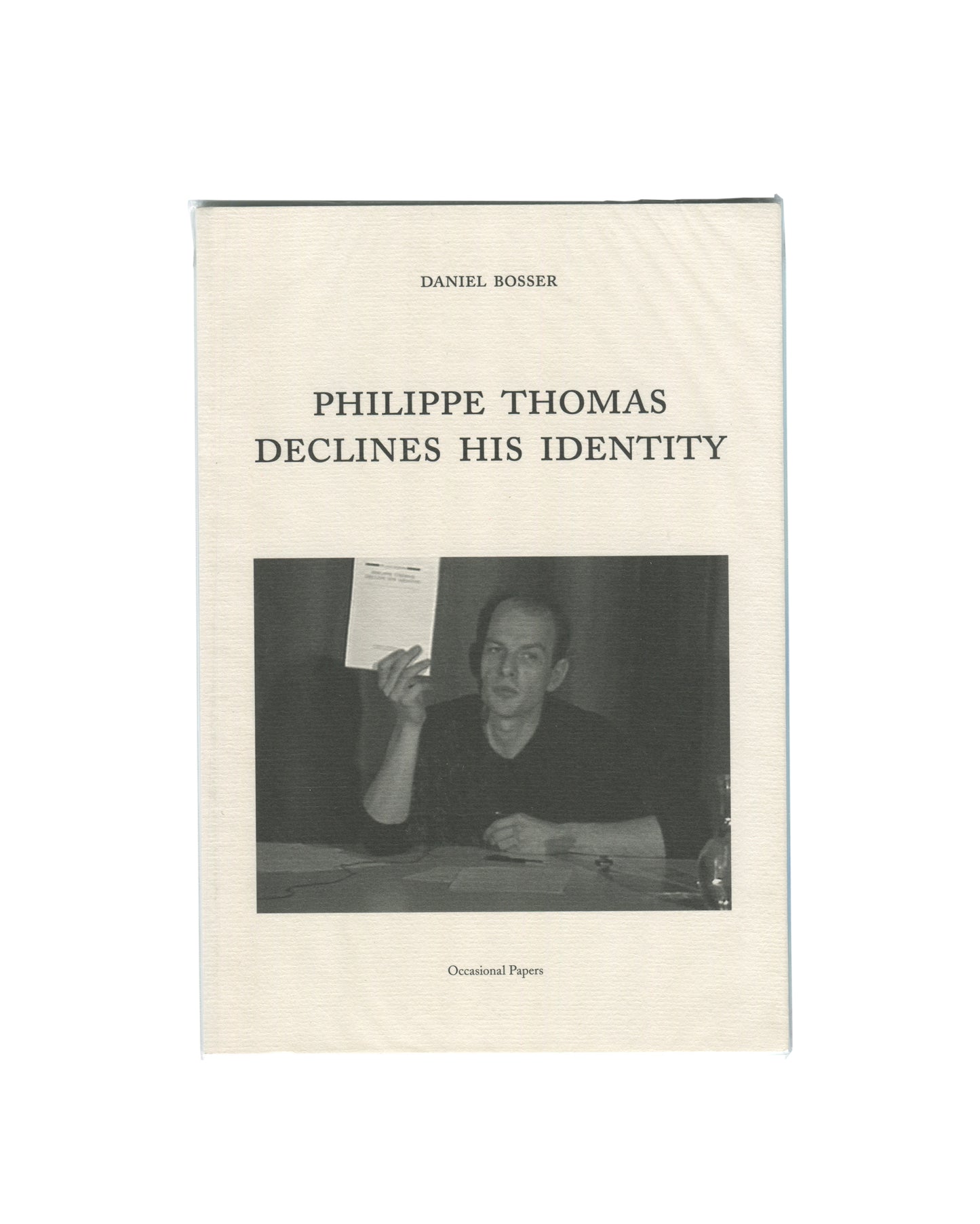 Original 2015 Daniel Bosser, Philippe Thomas Declines His Identity, Occasional Papers, London. Cream textured softcover with 'DANIEL BOSSER' in small spaced serif capitals at top, 'PHILIPPE THOMAS / DECLINES HIS IDENTITY' in larger spaced serif type at centre, and a black-and-white photograph below showing Philippe Thomas seated at a table, holding up a document, wearing a black top, expression direct and slightly confrontational. 'Occasional Papers' in italic type at foot.