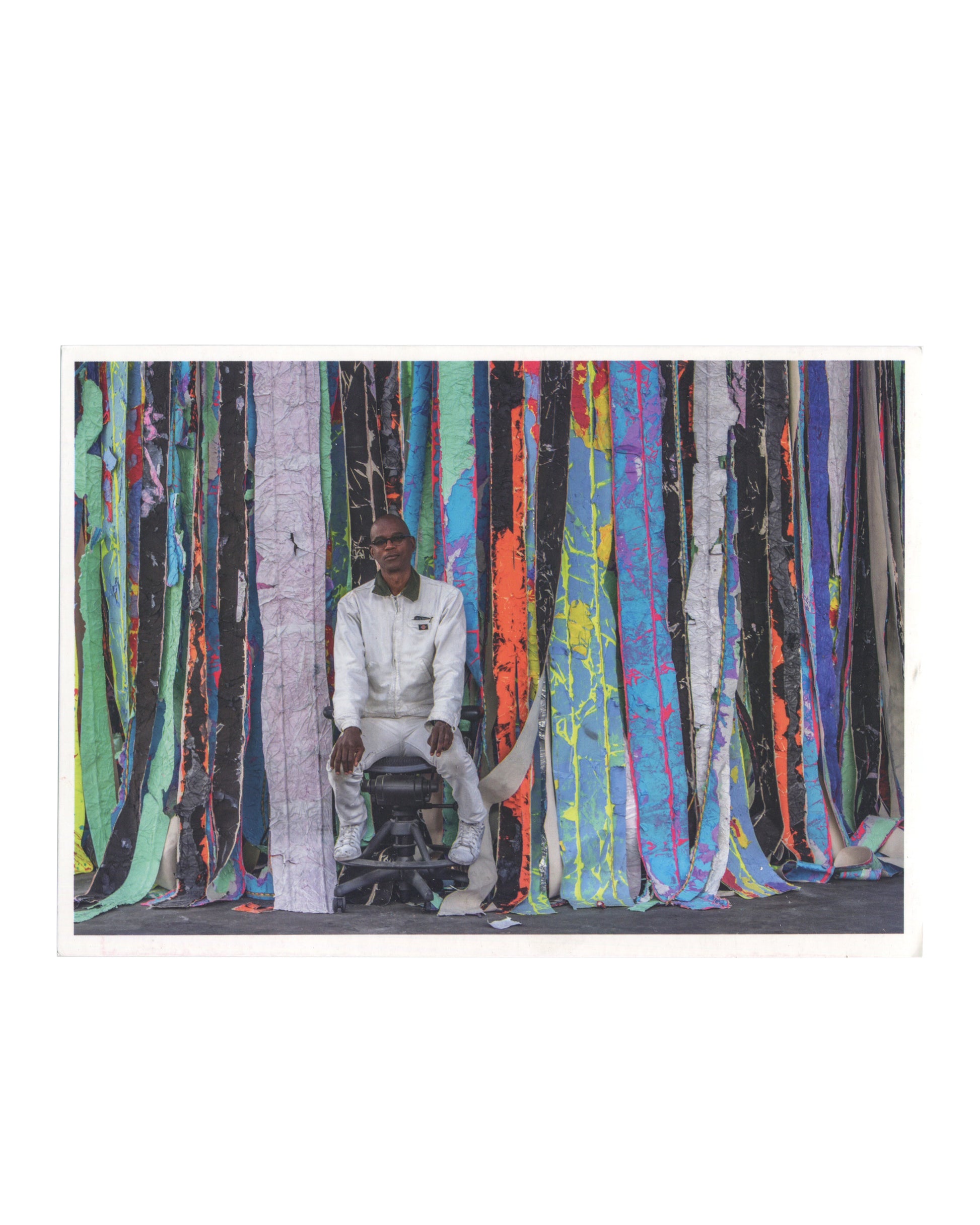 Reverse of 2019 Mark Bradford: Los Angeles exhibition card reproducing a photograph of the artist seated before Float (2019), a monumental installation of layered and stripped paper surfaces in vivid vertical bands of colour.