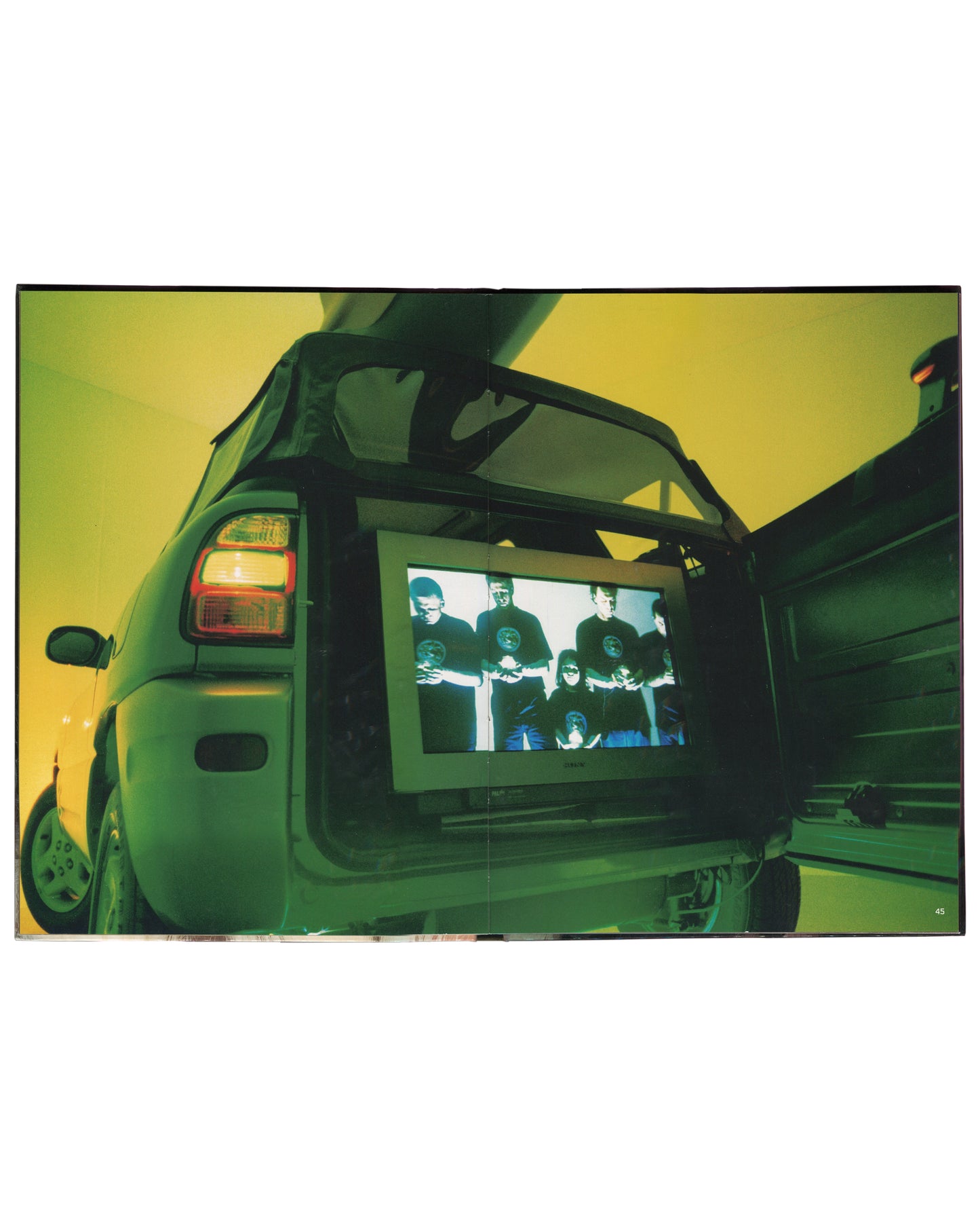 Interior spread of 2001 Olaf Breuning Ugly — full-bleed colour photograph of a large CRT television monitor mounted in the open boot of a hatchback car under a yellow-green sky, screen showing a group portrait of seven people in dark clothing