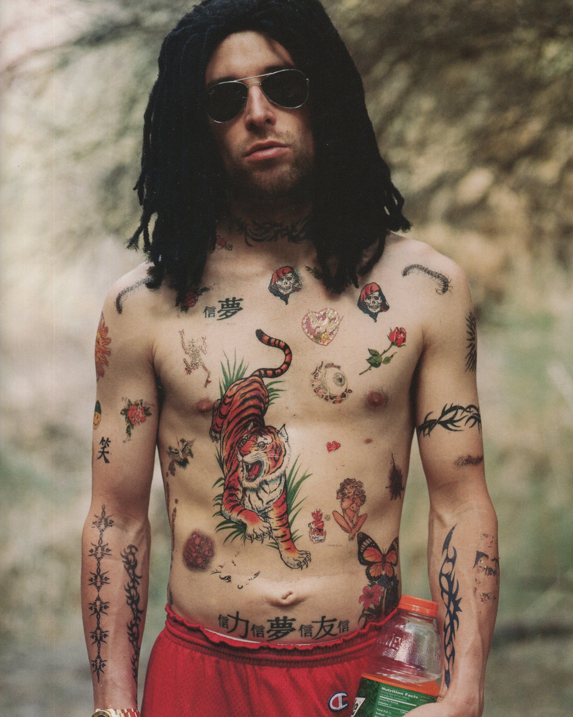 Interior spread of 2001 Olaf Breuning Ugly — full-page colour photograph of a young woman in round sunglasses, white crop top, and shoulder holster standing in a narrow wooden doorway, holding a pistol raised to shoulder and a green-and-red power drill at hip