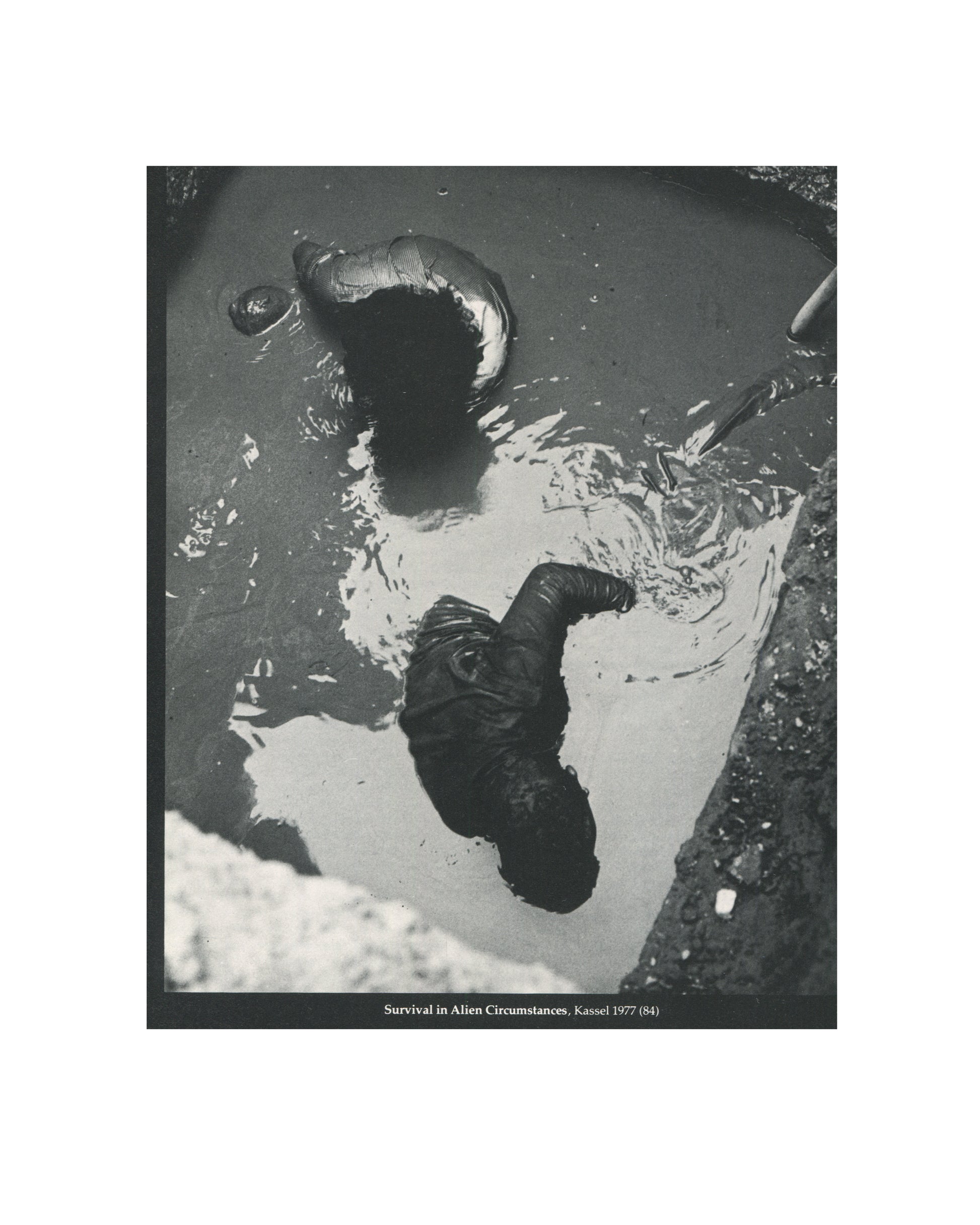 `Stuart Brisley — ICA, London, 1981 — interior plate. Black and white photograph documenting Survival in Alien Circumstances, Kassel, 1977. Aerial view of a figure submerged or lying in dark liquid, body contorted, clothing saturated, surrounded by debris and reflective water surface. Caption: "Survival in Alien Circumstances, Kassel 1977 (84)."`