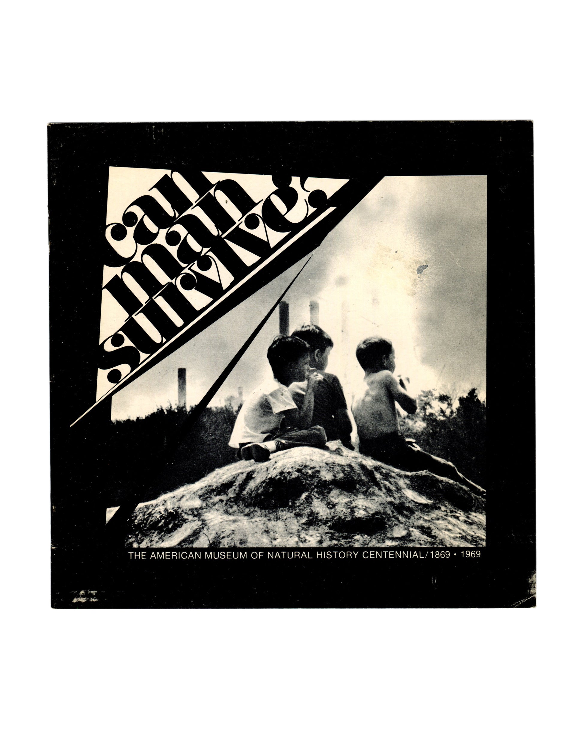  Original 1969 Can Man Survive? exhibition catalogue, American Museum of Natural History Centennial, New York. Front cover showing bold diagonal typographic design by Edward Krent — title text in black and white against a black ground — with a black-and-white photograph of three children seated on a mound of earth with industrial chimneys in the background. Staple-bound softcover, 20 × 20 cm.

