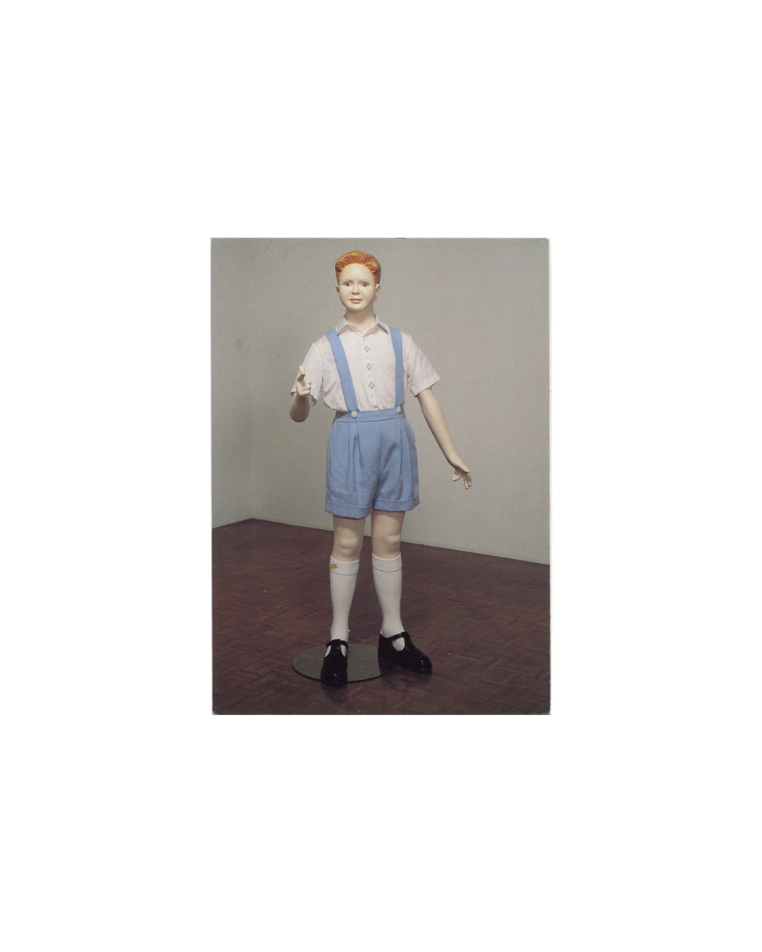 Postcard recto, Charles Ray, Boy, Whitney Museum of American Art, New York, 1997. Offset colour reproduction. Full-length view of sculpture — life-size figure of a boy with short red hair, pale complexion; white short-sleeve shirt with blue buttons, blue shorts with braces/suspenders, white knee socks, black Mary Jane shoes; standing on small black metal base against neutral grey gallery wall and dark wood floor. Painted fiberglass, steel, and fabric. Light surface marks, good vintage condition.

