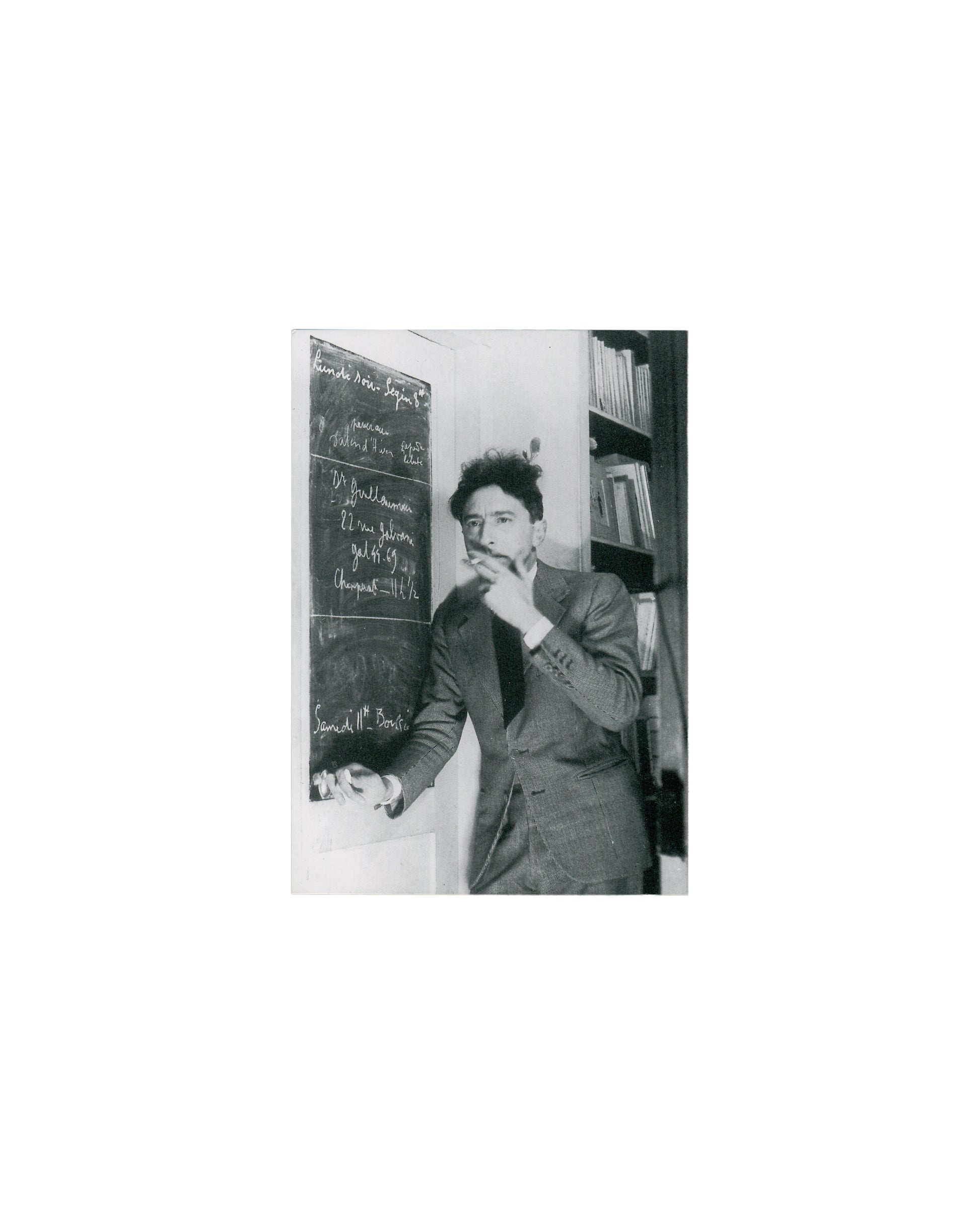 Recto — portrait Jean Cocteau, Poésie graphique, Galerie Arcade Colette, Paris, 1996. Invitation recto: monochrome photograph of Jean Cocteau — three-quarter view, standing beside a blackboard covered in chalk handwriting, right hand raised to chin, cigarette between fingers; dark suit and tie; bookshelves at right; chalk text partially legible including 'Lundi Noir' and names. Light age toning.