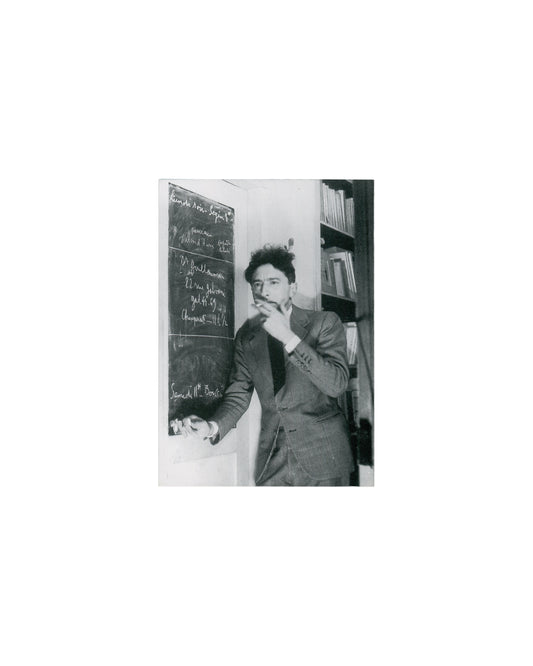  Recto — portrait Jean Cocteau, Poésie graphique, Galerie Arcade Colette, Paris, 1996. Invitation recto: monochrome photograph of Jean Cocteau — three-quarter view, standing beside a blackboard covered in chalk handwriting, right hand raised to chin, cigarette between fingers; dark suit and tie; bookshelves at right; chalk text partially legible including 'Lundi Noir' and names. Light age toning.