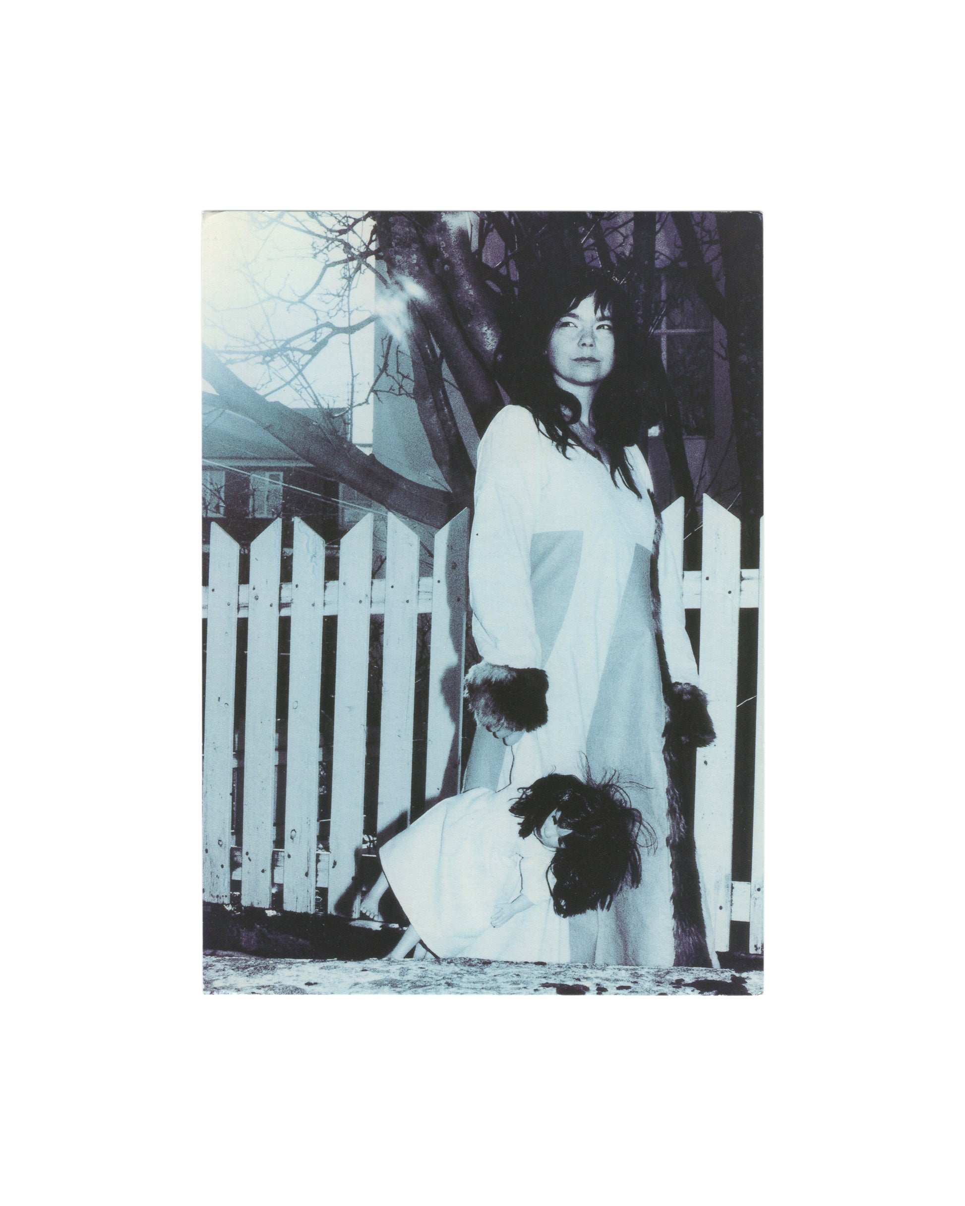 Anton Corbijn, Still Lives, exhibition opening card recto. Torch Gallery, Amsterdam, 2000. Blue-toned black-and-white photograph: a woman with long dark hair stands in a white dress beside a white picket fence, bare winter trees behind her. A second figure in a matching white dress crouches at her feet, face obscured by dark hair. Atmospheric, gothic mood. Image: Anton Corbijn, Björk, Reykjavik, 1999.
