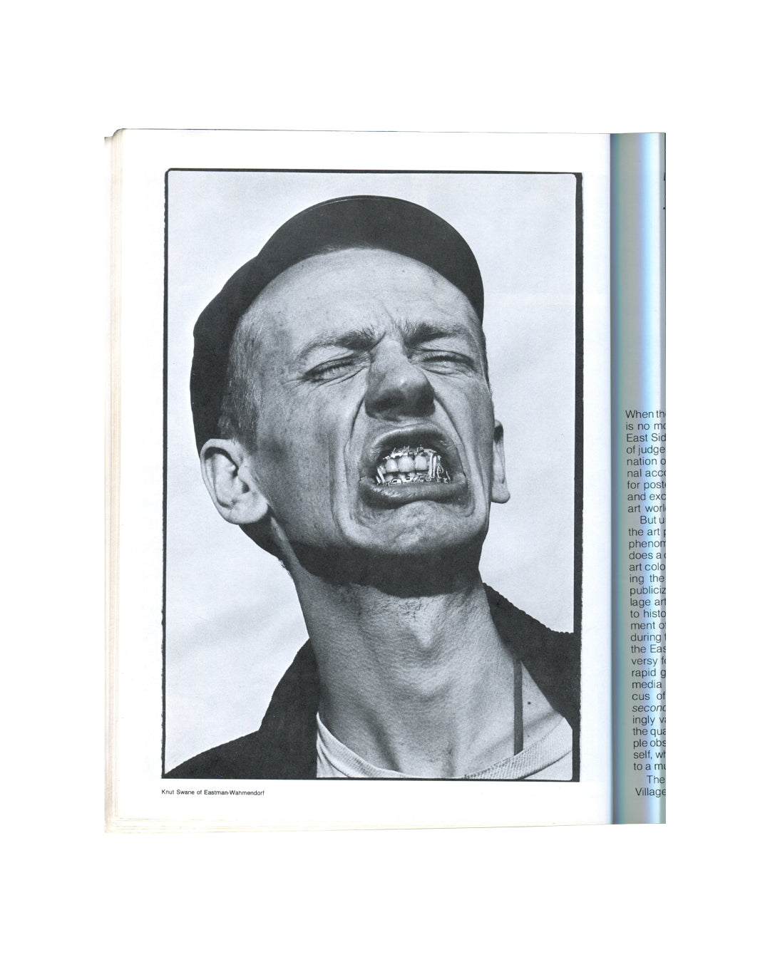East Village '85: A Guide. A Documentary, Pelham Press, 1985. Interior spread: essay opening. Left: full-page black-and-white portrait photograph — Knut Swane of Eastman-Wahmendorf, mouth open wide in an expression of anguish or ecstasy, gold teeth visible, head tilted back, wearing a dark jacket. Right: essay text column beginning 'When th[e]...' discussing the East Village art scene, media attention, and the conditions of downtown New York art life. Downtown New York, 1980s art scene.
