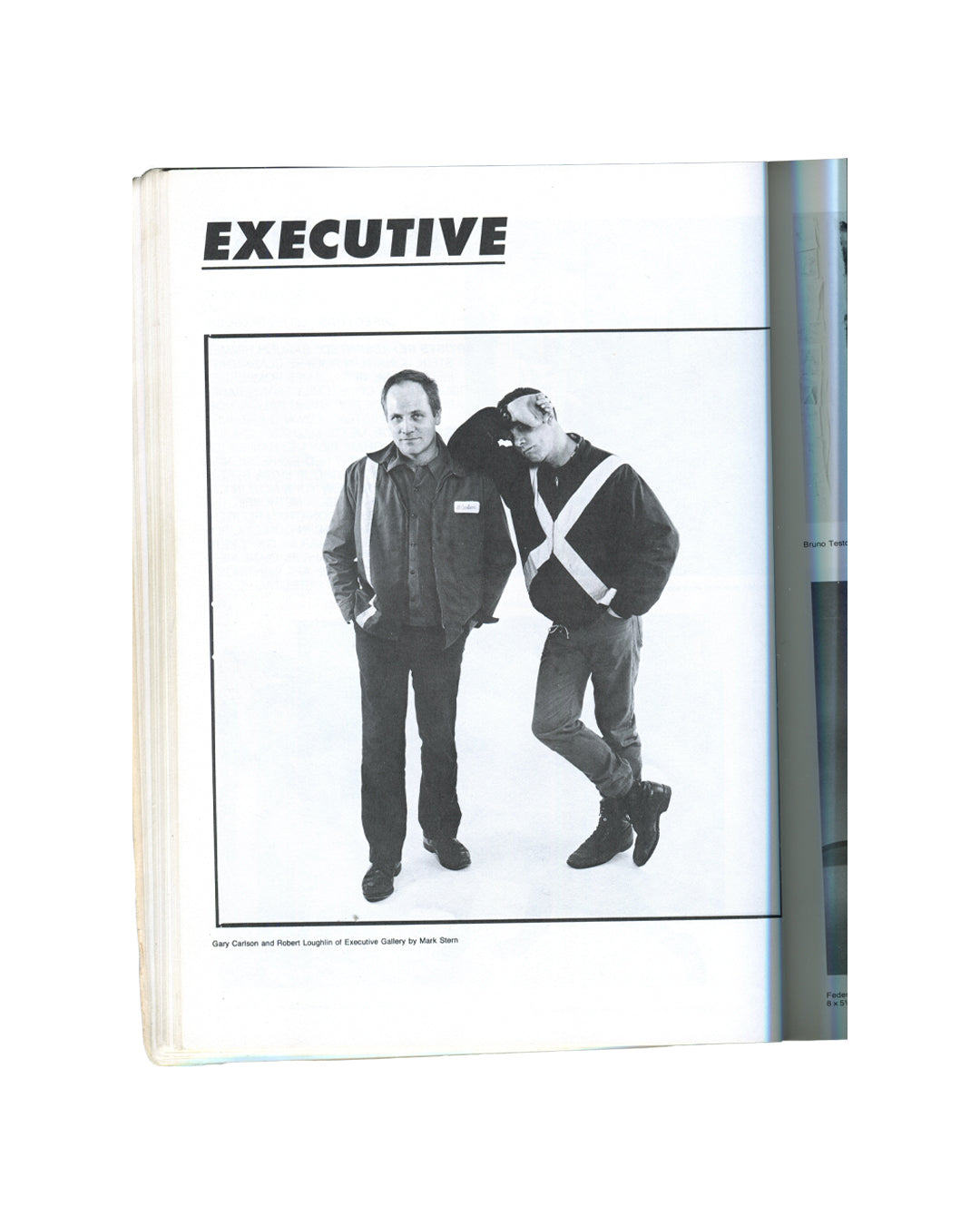 East Village '85: A Guide. A Documentary, 1985. Interior page: Executive Gallery listing. Bold title 'EXECUTIVE'. Black-and-white photograph by Mark Stern: Gary Carson and Robert Loughlin of Executive Gallery — two men standing against a white ground, one in a dark jacket with a name badge, the other in a black top with a large white X across the chest, leaning into him casually. East Village gallery directory.
