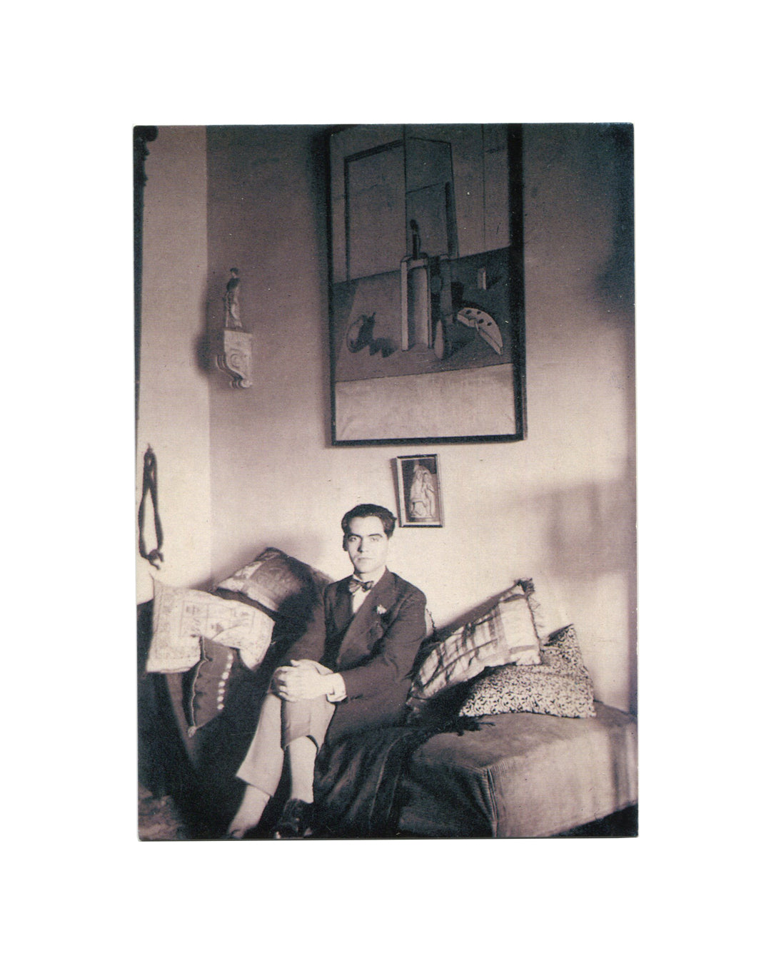 Fundación Federico García Lorca postcard, c.1998. Recto: black-and-white photograph of Federico García Lorca seated on a sofa in the family home, Granada, 1925, wearing a dark suit and bow tie, hands clasped; above him on the wall, a Salvador Dalí still life painting showing bottles and objects on a ledge; small devotional image below the painting; cushions and domestic objects visible around him.