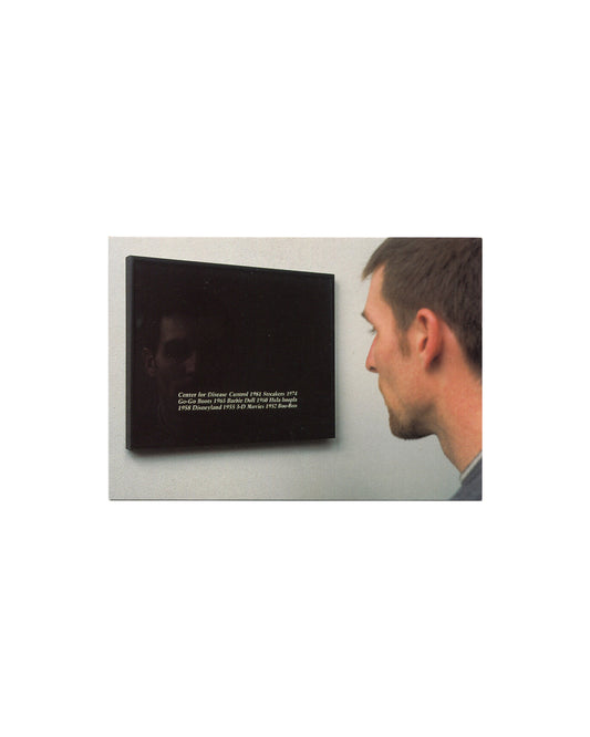 Original 2000 Felix Gonzalez-Torres Serpentine Gallery exhibition postcard, London. Recto showing installation photograph of "Untitled", 1988, framed photostat — a young man in profile reading white text on a black surface reading Center for Disease Control 1981 Streakers 1974 Go-Go Boots 1965 Barbie Doll 1960 Hula hoopla 1958 Disneyland 1955 3-D Movies 1952 Boo-Boo, his faint reflection visible in the dark surface.