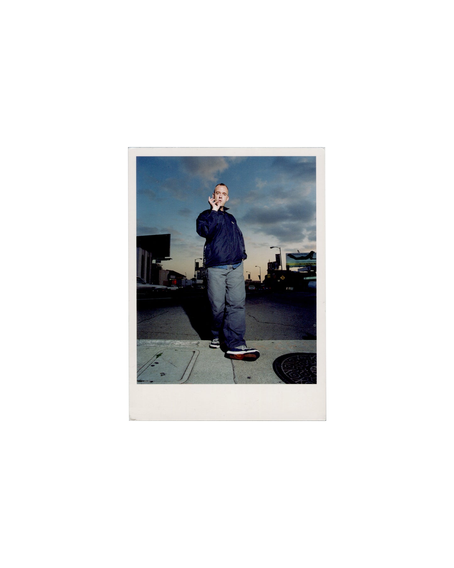 Recto Martin Gardiner, Norman Cook (Fatboy Slim), Los Angeles, 1999. NPG postcard, 2001. Recto: colour photograph — full-length portrait of a shaven-headed man in a dark zip-up jacket and grey trousers, standing on a Los Angeles pavement, right hand raised to mouth, cigarette between fingers; dramatic cloudy sky at dusk; street furniture and parked vehicles in background. Polaroid-style white border