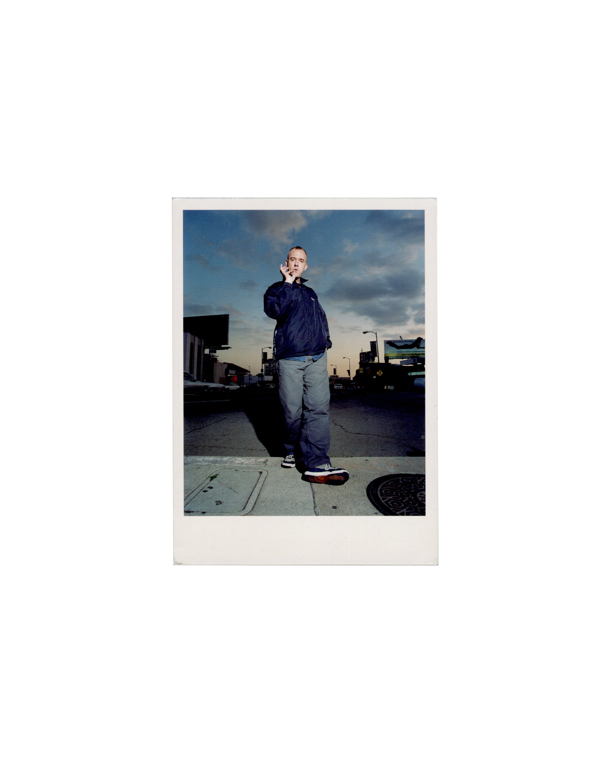 Recto Martin Gardiner, Norman Cook (Fatboy Slim), Los Angeles, 1999. NPG postcard, 2001. Recto: colour photograph — full-length portrait of a shaven-headed man in a dark zip-up jacket and grey trousers, standing on a Los Angeles pavement, right hand raised to mouth, cigarette between fingers; dramatic cloudy sky at dusk; street furniture and parked vehicles in background. Polaroid-style white border
