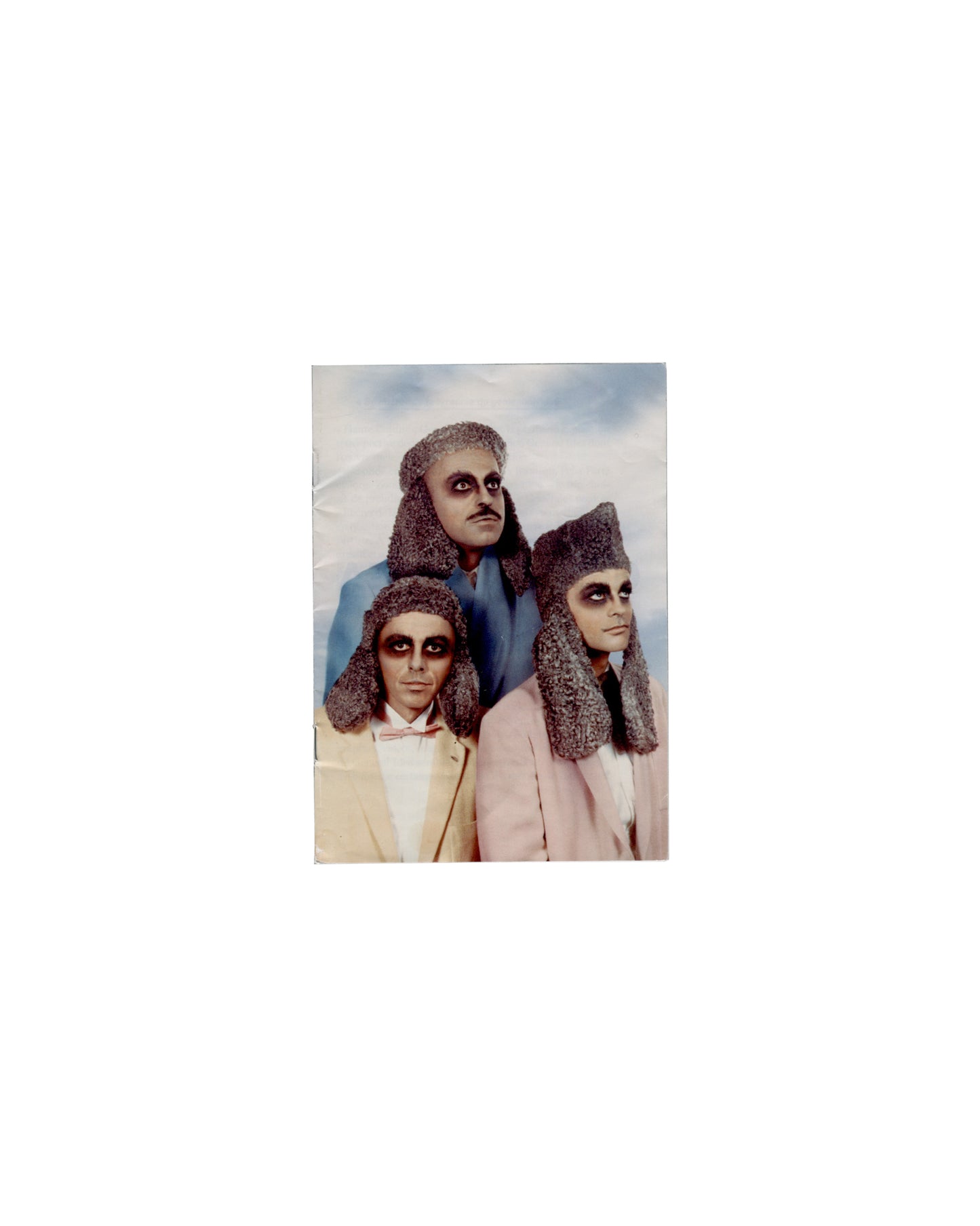 Front cover General Idea, Haute Culture: Une rétrospective, 1969–1994, Musée d'Art moderne de la Ville de Paris, 2011. Booklet front cover: colour photograph of three figures in theatrical costume — heavy dark eye makeup, textured grey headdresses, pastel suits (cream, blue, pink); pyramid composition against pale blue-grey sky; staged, deadpan. Handling creases, surface undulation.