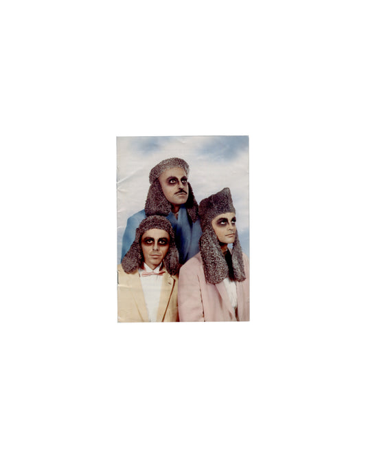 Front cover General Idea, Haute Culture: Une rétrospective, 1969–1994, Musée d'Art moderne de la Ville de Paris, 2011. Booklet front cover: colour photograph of three figures in theatrical costume — heavy dark eye makeup, textured grey headdresses, pastel suits (cream, blue, pink); pyramid composition against pale blue-grey sky; staged, deadpan. Handling creases, surface undulation.