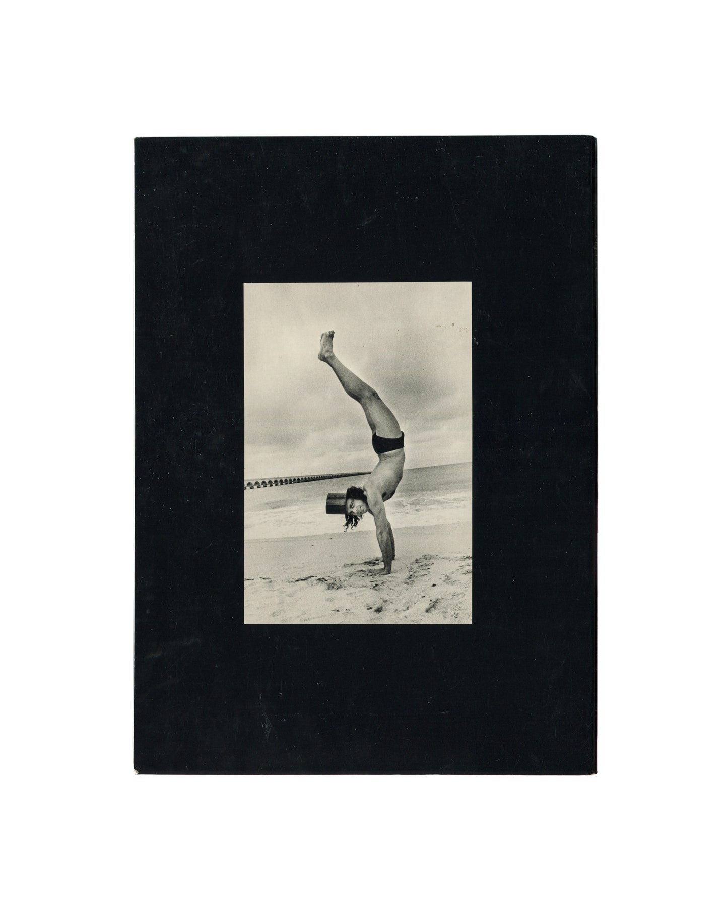 Reverse of Ralph Gibson, Deja-Vu, Lustrum Press, 1973, showing a black-and-white photograph of a figure performing a one-armed handstand on a beach in swimwear and a top hat, mounted on black card.