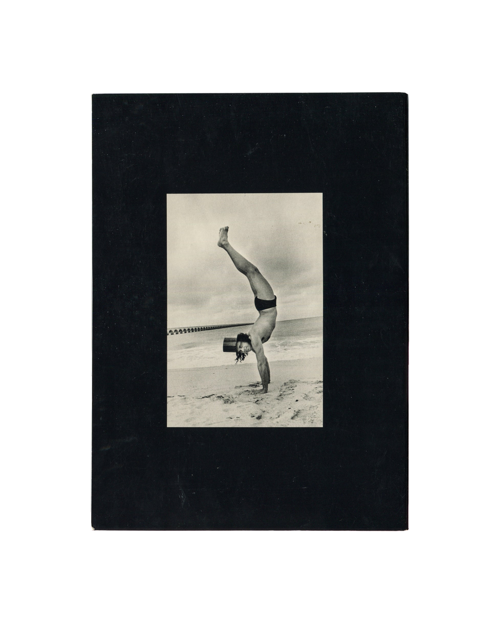 Reverse of Ralph Gibson, Deja-Vu, Lustrum Press, 1973, showing a black-and-white photograph of a figure performing a one-armed handstand on a beach in swimwear and a top hat, mounted on black card.