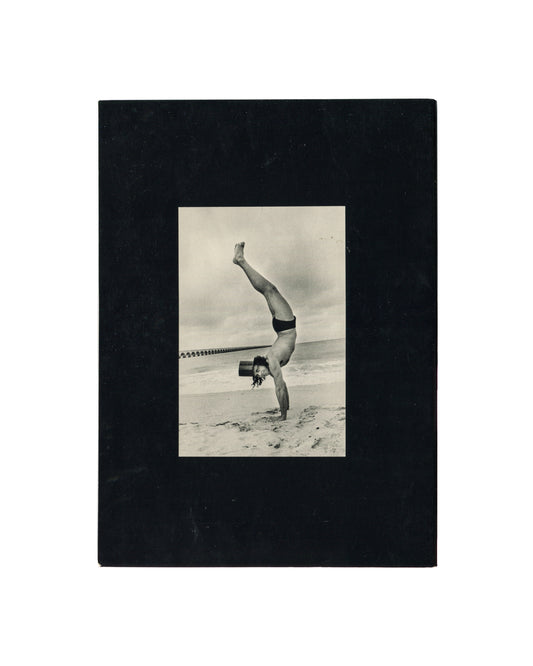 Reverse of Ralph Gibson, Deja-Vu, Lustrum Press, 1973, showing a black-and-white photograph of a figure performing a one-armed handstand on a beach in swimwear and a top hat, mounted on black card.