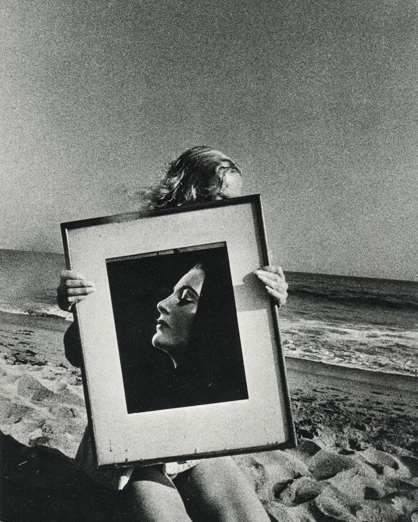  Interior page of Ralph Gibson, Deja-Vu, Lustrum Press, 1973, showing a figure on a beach holding a framed black-and-white portrait photograph of a woman in profile before their own face, sea and sky visible behind.