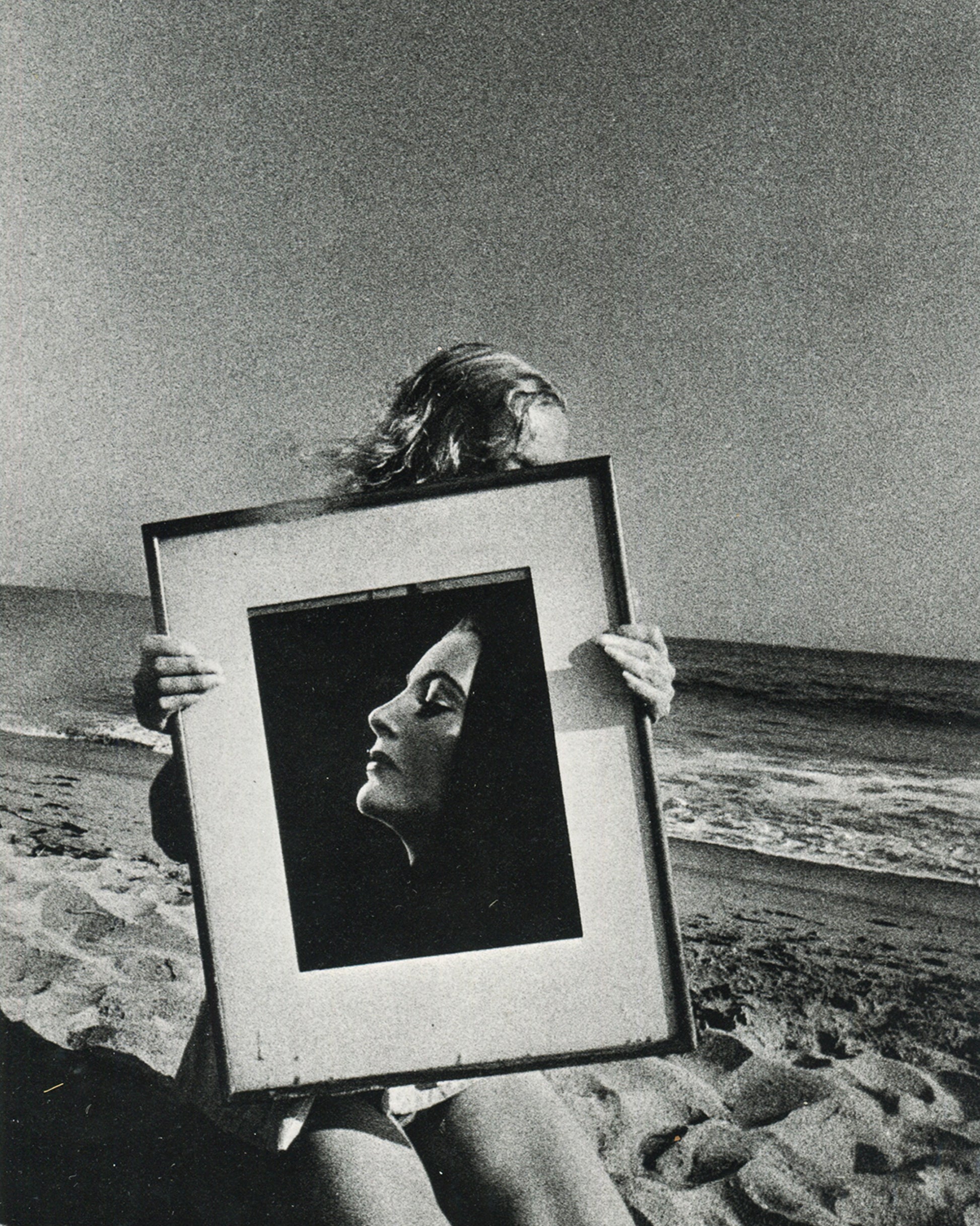  Interior page of Ralph Gibson, Deja-Vu, Lustrum Press, 1973, showing a figure on a beach holding a framed black-and-white portrait photograph of a woman in profile before their own face, sea and sky visible behind.