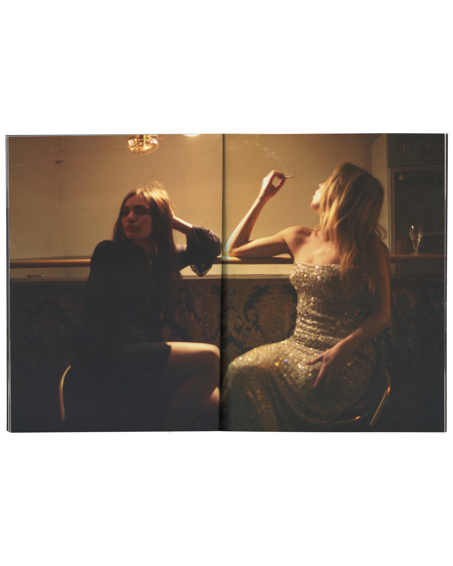 Interior spread from 2017 Nan Goldin, Biesenthal. Full-bleed double-page colour photograph of two women in a warmly lit interior — left figure in dark clothing, sunglasses, resting head on hand; right figure in a silver sequined strapless dress, arm raised, cigarette in hand. A chandelier glows above, a wine glass visible at right. Amber light, smoke, and soft focus throughout.