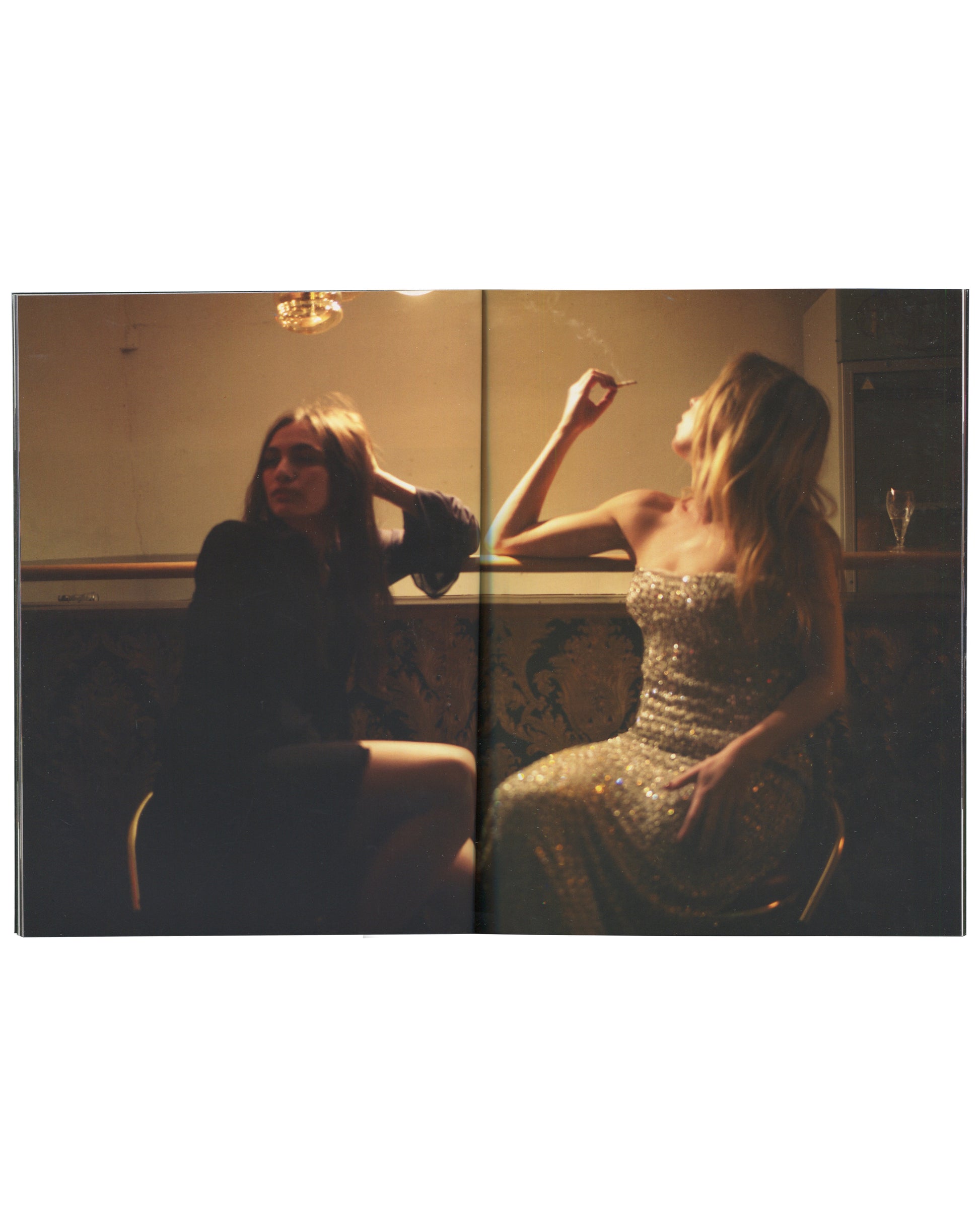 Interior spread from 2017 Nan Goldin, Biesenthal. Full-bleed double-page colour photograph of two women in a warmly lit interior — left figure in dark clothing, sunglasses, resting head on hand; right figure in a silver sequined strapless dress, arm raised, cigarette in hand. A chandelier glows above, a wine glass visible at right. Amber light, smoke, and soft focus throughout.