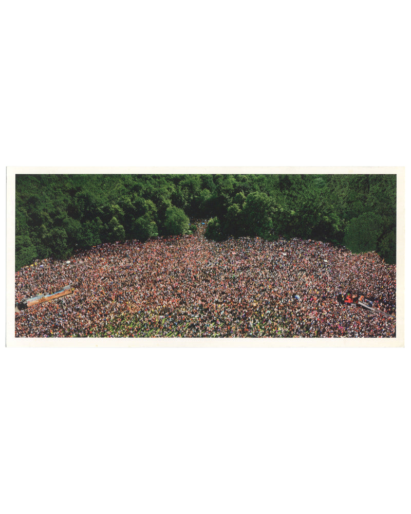 Andreas Gursky, Love Parade, 2001, artist postcard recto. Matthew Marks Gallery, New York, 2001. Colour reproduction of a large-format C-print: aerial view of an enormous crowd filling a clearing in a dense green forest, photographed from directly above. The mass of people — hundreds of thousands — forms an irregular oval shape, their individual figures indistinguishable at this scale, creating a dense texture of skin tones, clothing, and colour. Two long vehicles or floats are visible at the left and lower