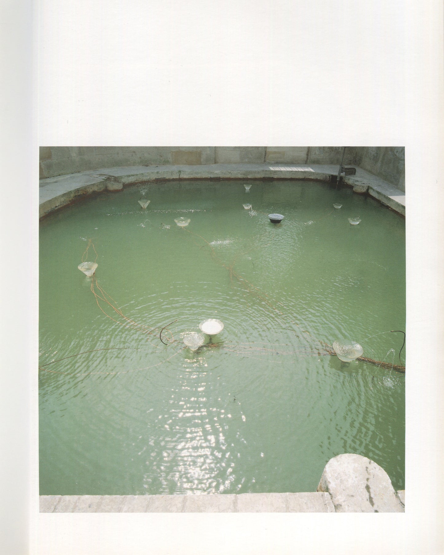 Interior page from Rebecca Horn Missing Full Moon, Artsite Gallery, Bath, 1989. Overhead photograph of a large circular stone basin filled with green-tinted water, with multiple small glass funnel forms floating on the surface connected by fine wires radiating from the centre.