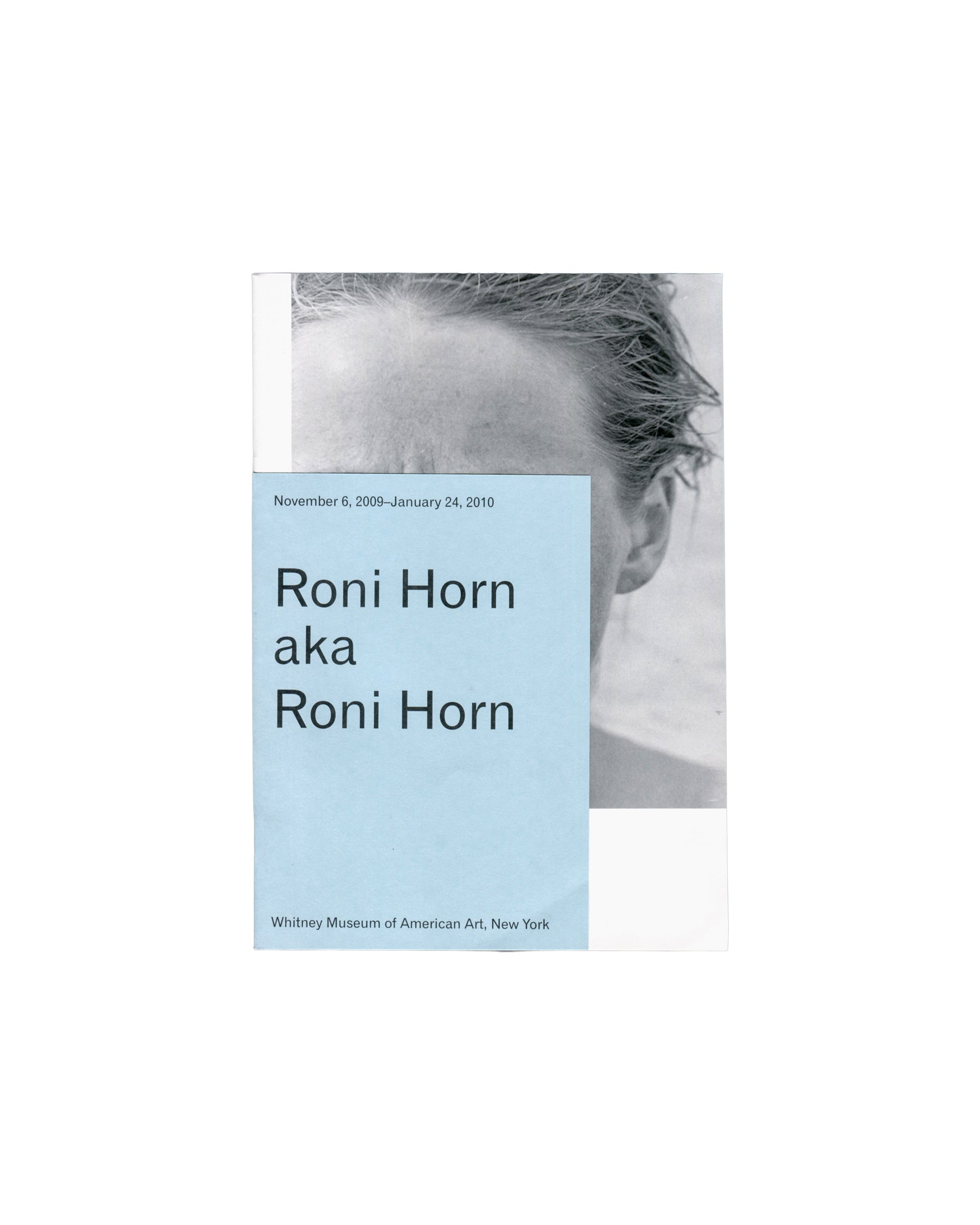 Front cover — black-and-white portrait with overwrap Roni Horn aka Roni Horn, Whitney Museum of American Art, New York, 2009. Exhibition guide front cover: pale blue printed overwrap overlaid on self-wrapper; overwrap carries "November 6, 2009–January 24, 2010" in small dark grey sans-serif type upper left; "Roni Horn / aka / Roni Horn" in large dark grey sans-serif type centre left; "Whitney Museum of American Art, New York" in small dark grey sans-serif type lower left; behind overwrap, black-and-white cl